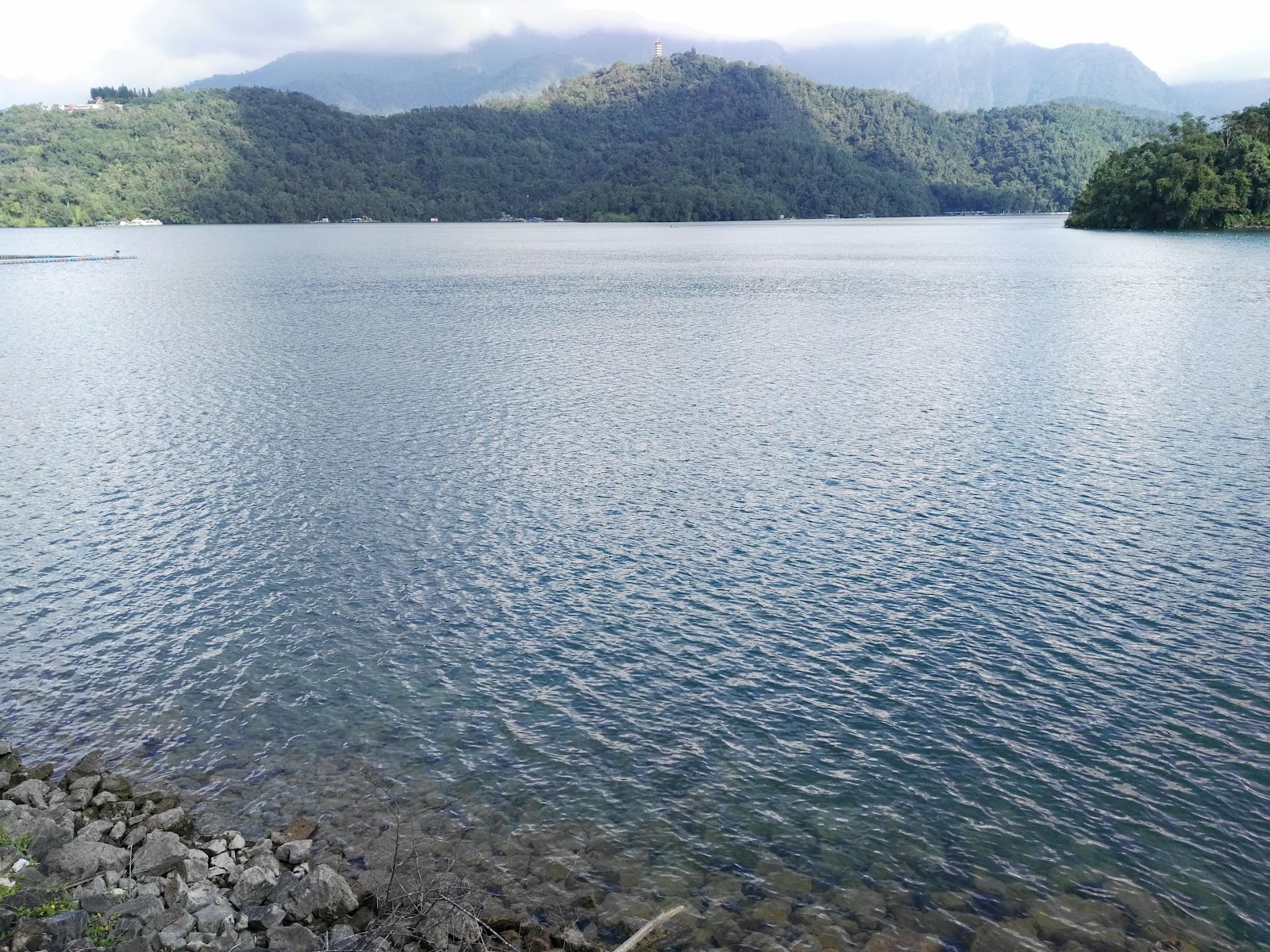 向山岸邊礁石近景，湖水清澈波光粼粼，遠山薄霧中若隱若現