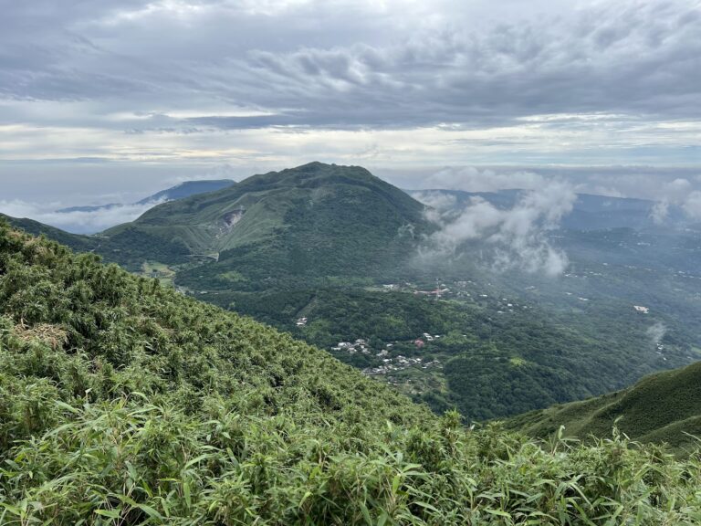 陽明山山頂俯瞰視角，層巒疊嶂綠意，遠方山谷聚落雲霧繚繞