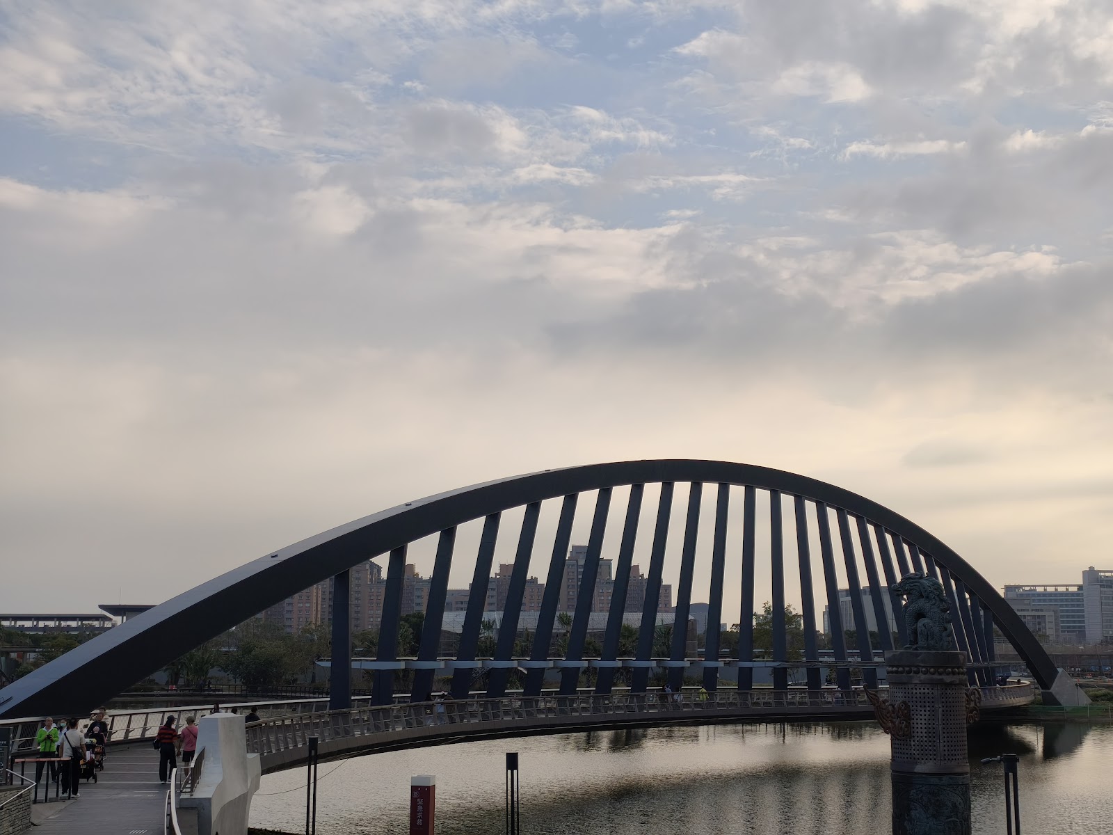 故宮南院寬月橋黑色拱橋，遊客在橋上行走，湖面映天，背景嘉義市景