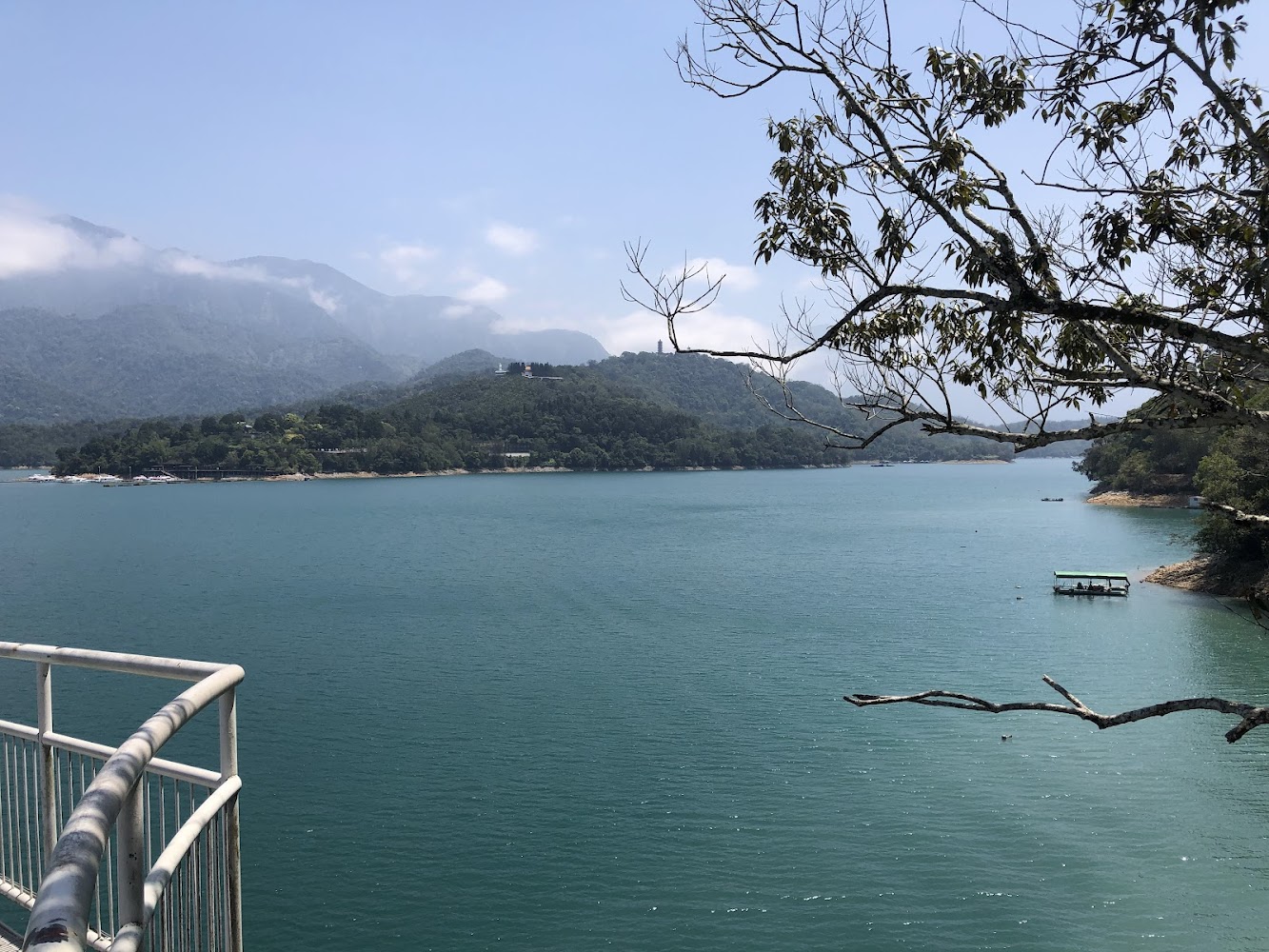 這張圖片主要呈現一片碧綠的湖,湖岸被青山環繞,遠方有雲霧繚繞的山巒。右側有一棵樹枝延伸,湖面上有一艘小船,前方則可見欄杆。圖片無浮水印。