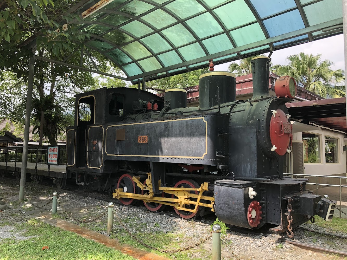 這張圖片展示了一輛復古蒸汽火車頭,編號366,位於室外並有透明棚子遮蔽,四周有樹木和建築。畫面左側有警告標示,並未見浮水印。
