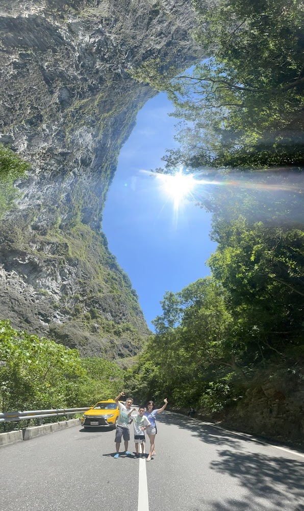 高聳山谷道路旁停著黃色汽車，三人合影，陽光灑落在峽谷間