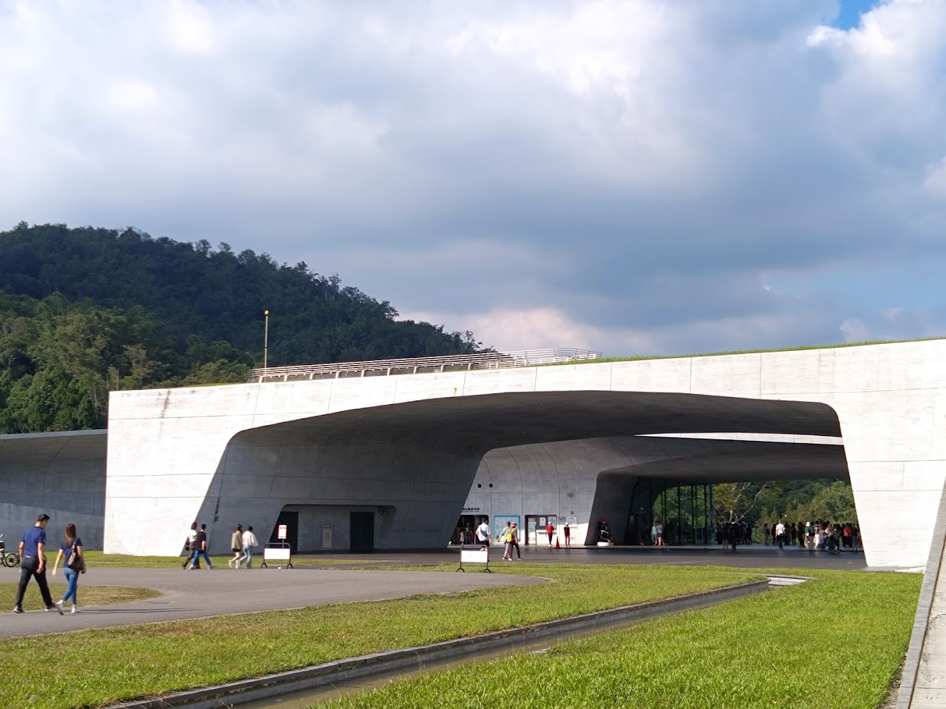 這張圖片主要是一座現代風格的建築,外觀為大型白色混凝土結構,周圍有綠地和遠處的樹林,有許多人走動或參觀。圖片中未見明顯浮水印。