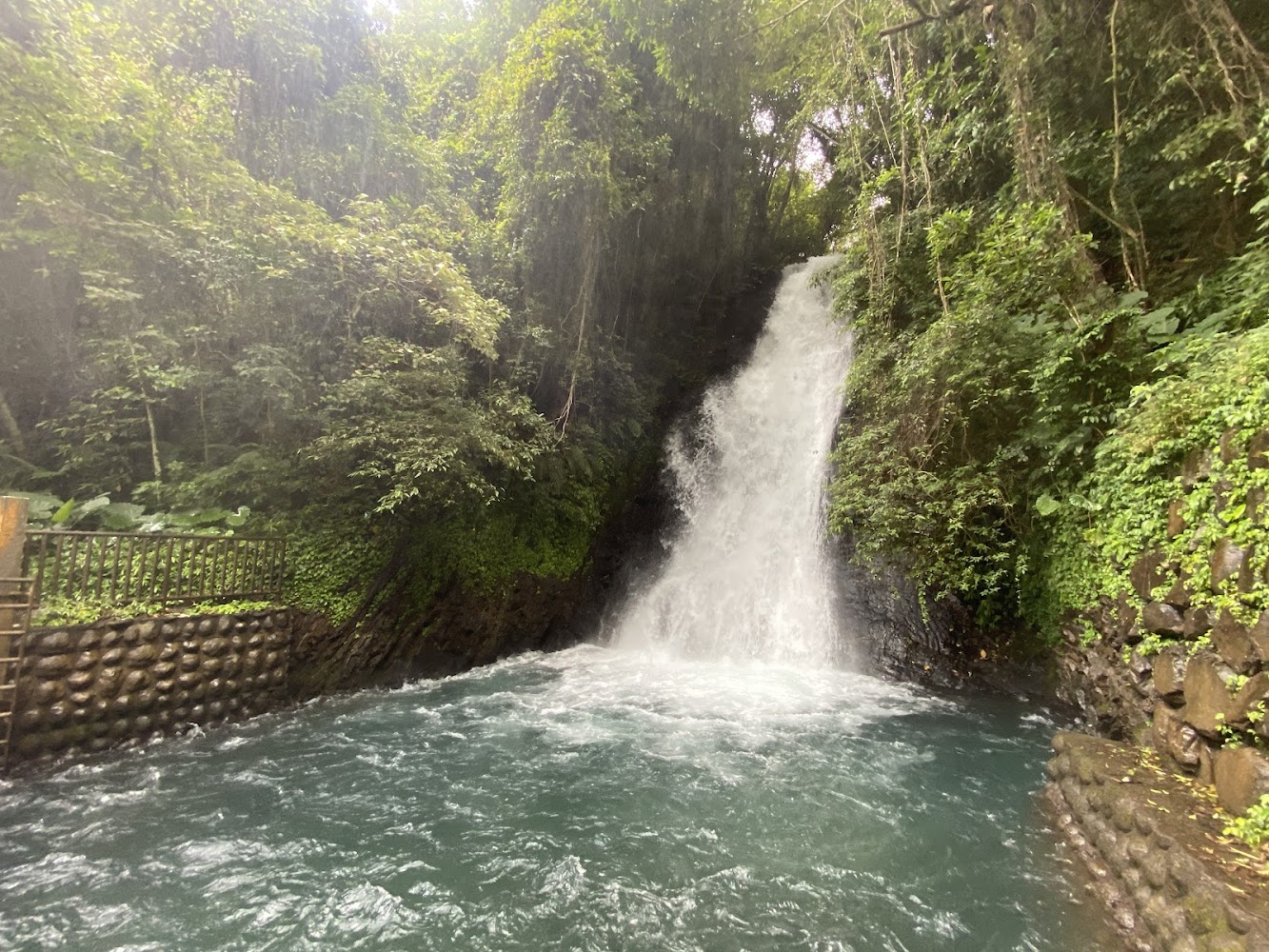 這張圖片以瀑布為主景,水流從高處傾瀉而下,匯聚成一個碧藍色的水池,四周被茂密的綠樹和石牆包圍。圖片未見明顯浮水印。
