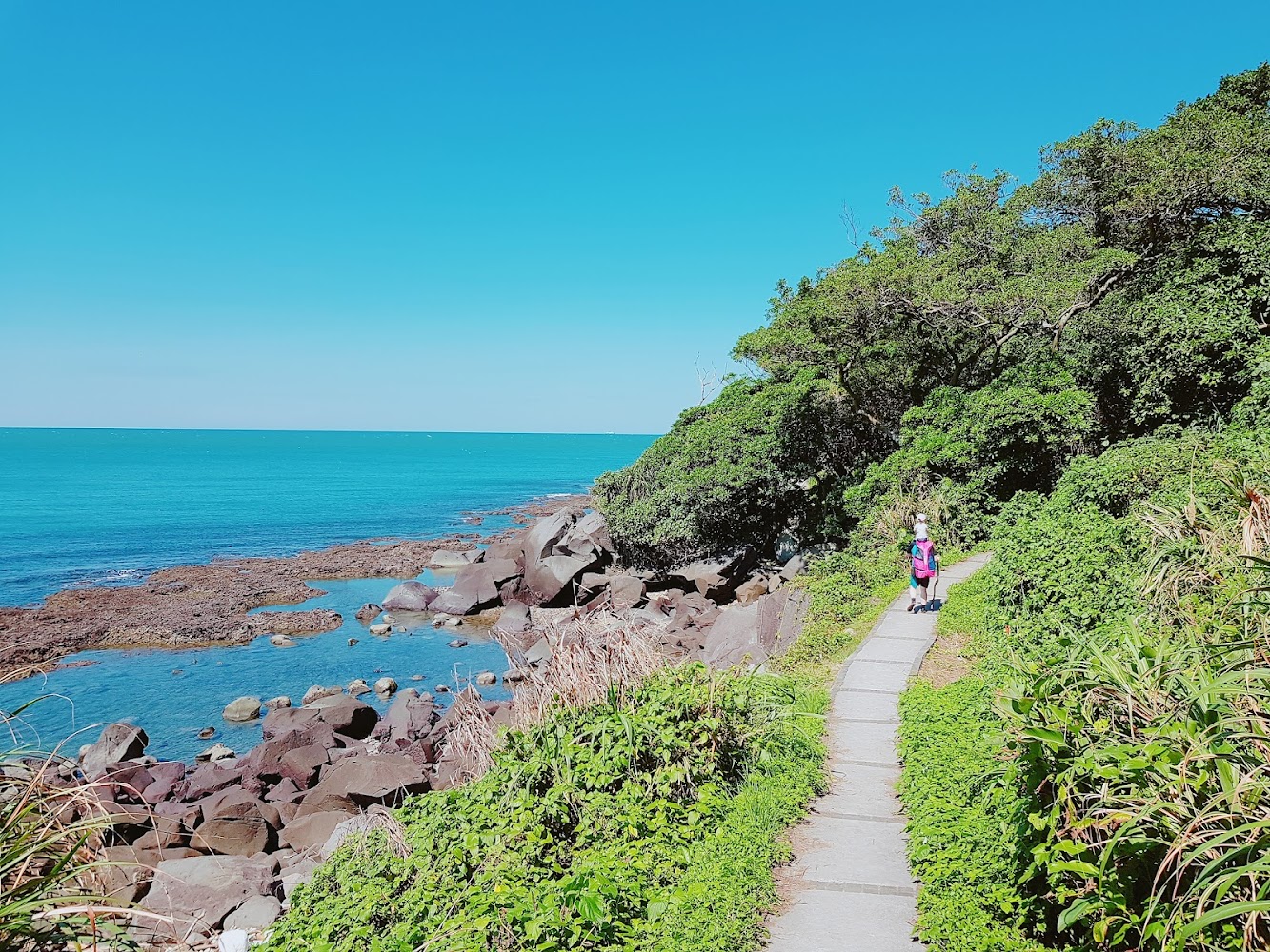 這張圖片展示蔚藍的海岸線、一條沿著海邊蜿蜒的步道,旁有翠綠茂密的植被和礁石,畫面中有人正在步行於步道上。圖片未見明顯浮水印。