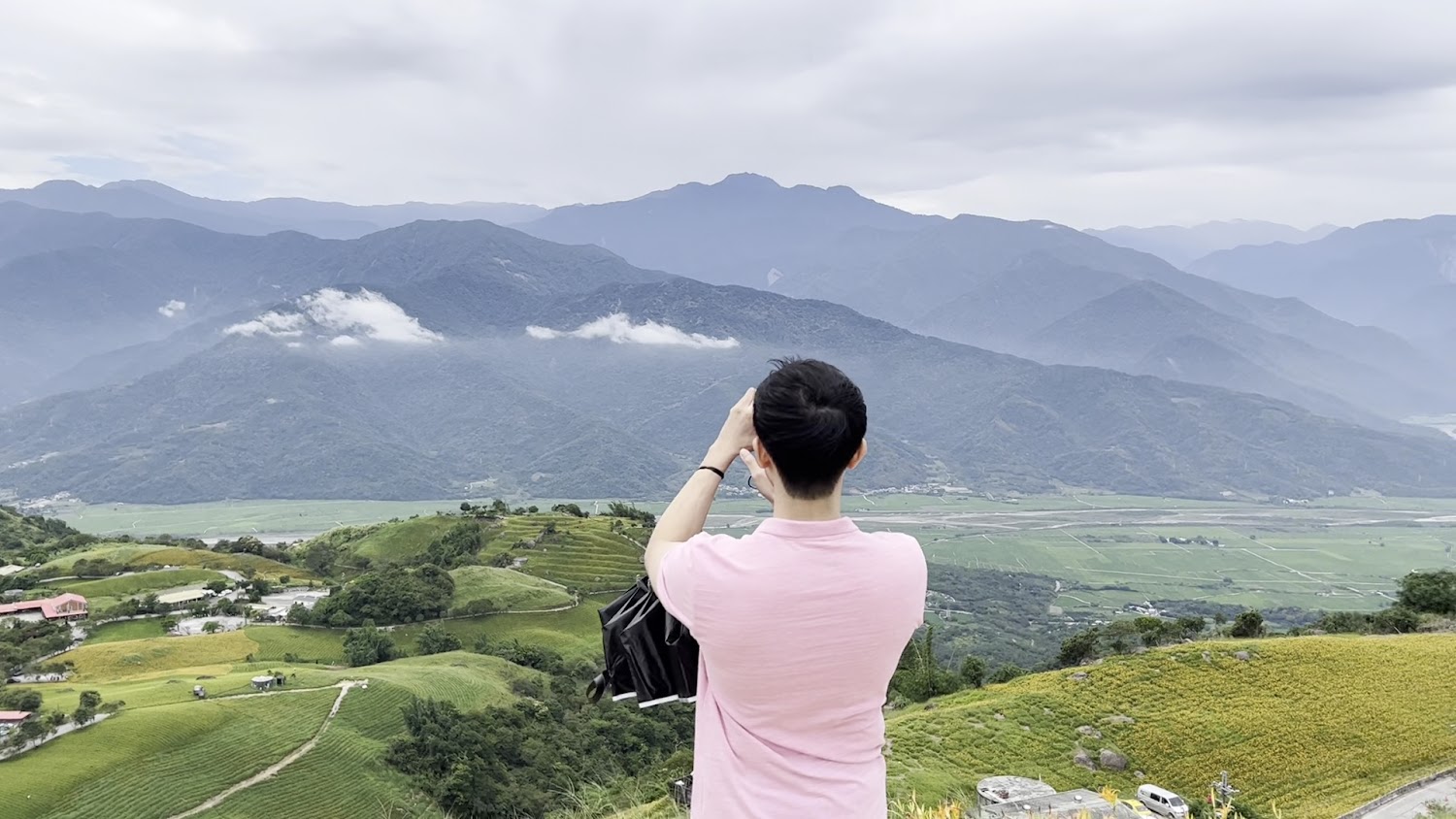 旅人站在山上遠眺層層梯田、綠丘與遠方山脈雲霧