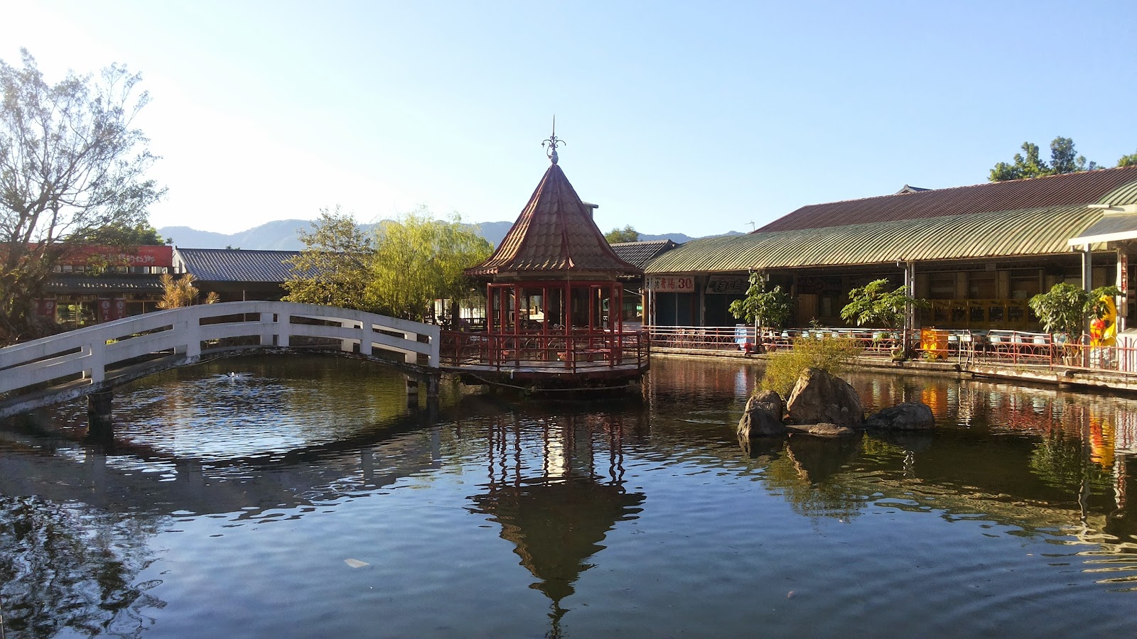花蓮觀光糖廠紅色六角涼亭與白橋水池全景，背景有山景與商店