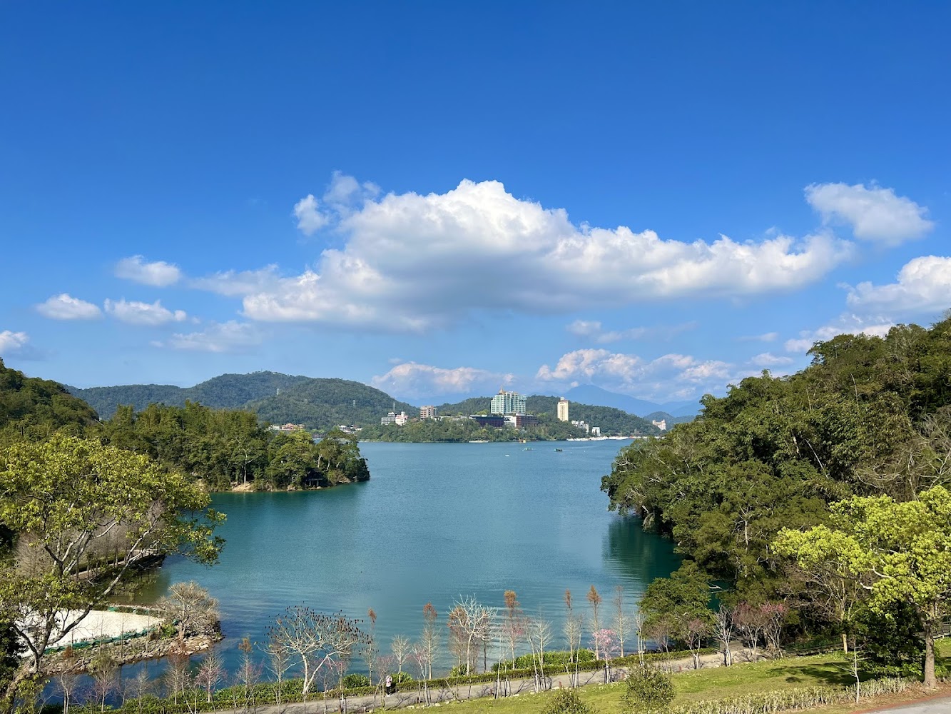 這張圖片主要展現一片寬闊的湖泊,湖水清澈,四周環繞著綠樹與山巒,湖邊有道路和植栽,遠處則可見一些建築物和藍天白雲。圖片無明顯浮水印。