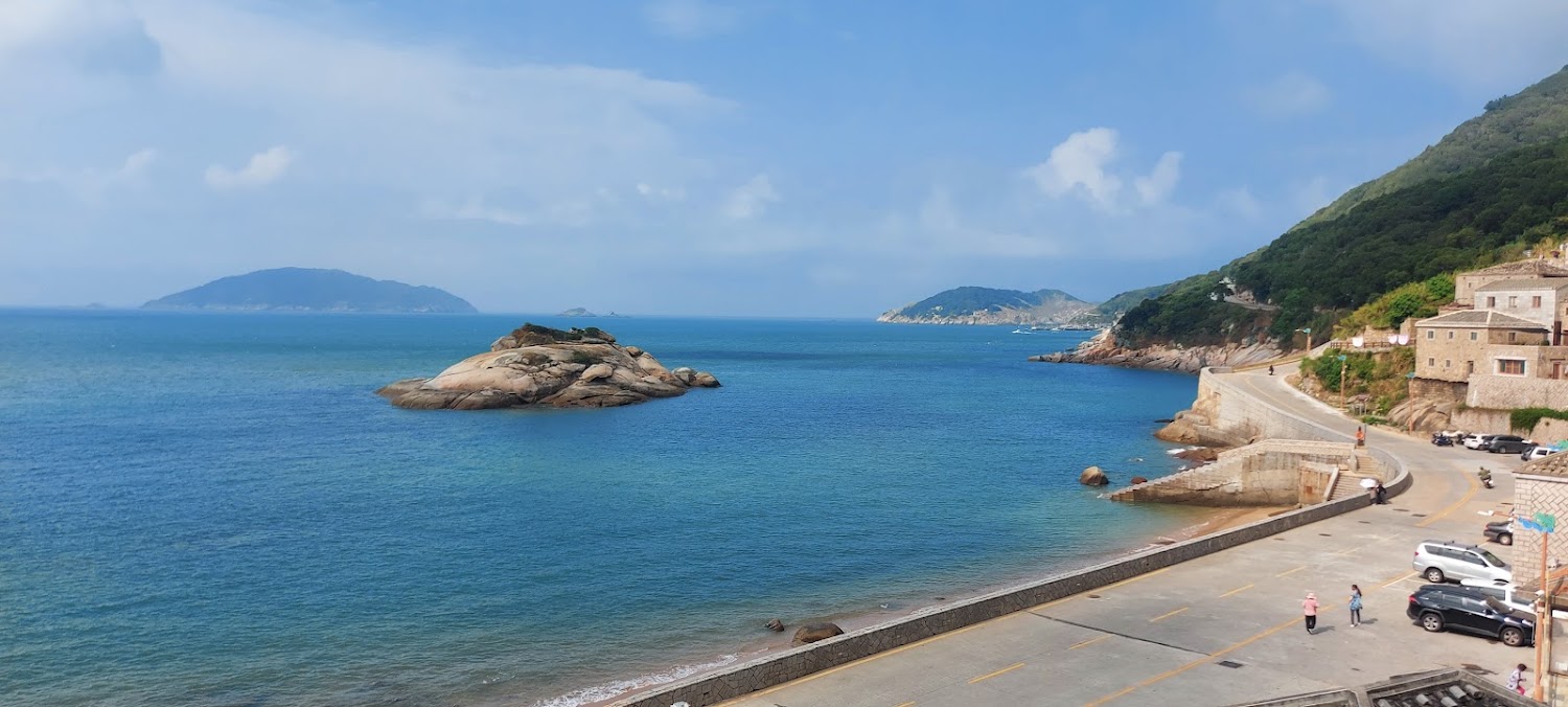 蔚藍海洋與礁石小島，右側村莊沿著海邊道路展開