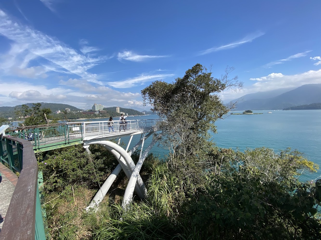這張圖片主要展示一座跨越湖畔的觀景平台,旁邊有綠樹與蔚藍湖水,遠處可見山脈與建築物。天空晴朗,沒有浮水印。