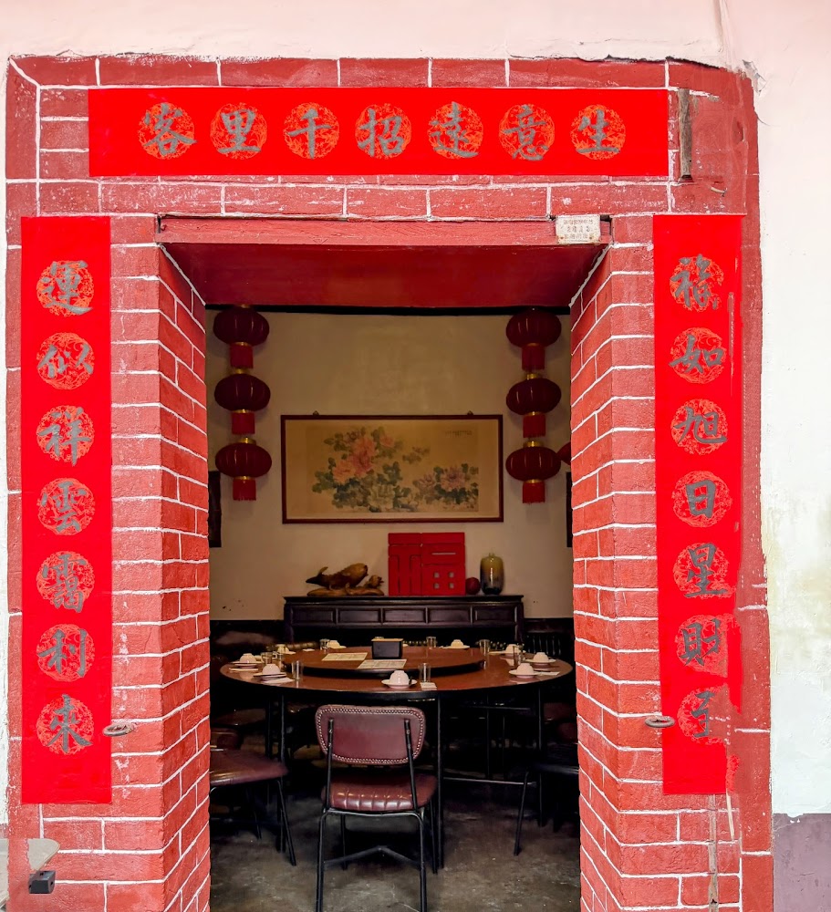 這張圖片展示了一間具中國傳統風格的餐廳入口，紅磚拱門上貼有紅色春聯與圓形字樣，門內有圓桌、餐具、掛畫和紅燈籠，營造出溫馨喜慶的中式氛圍，圖片無明顯浮水印。