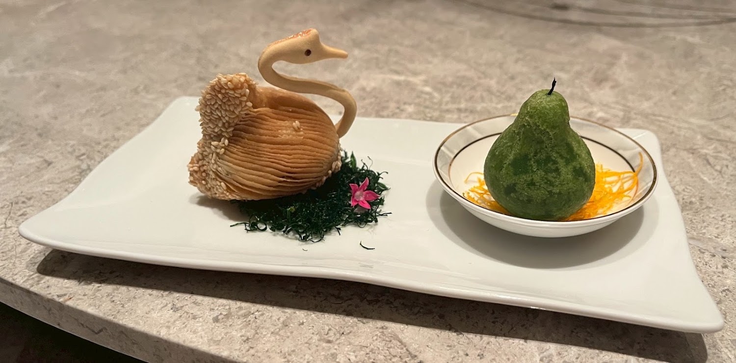 這張圖片展示了兩個精緻的中式點心，一個造型像天鵝，裝飾有芝麻與小花草，另一個則是綠色果型點心，底下襯著蛋絲，擺放於白色瓷盤。圖片中無浮水印。