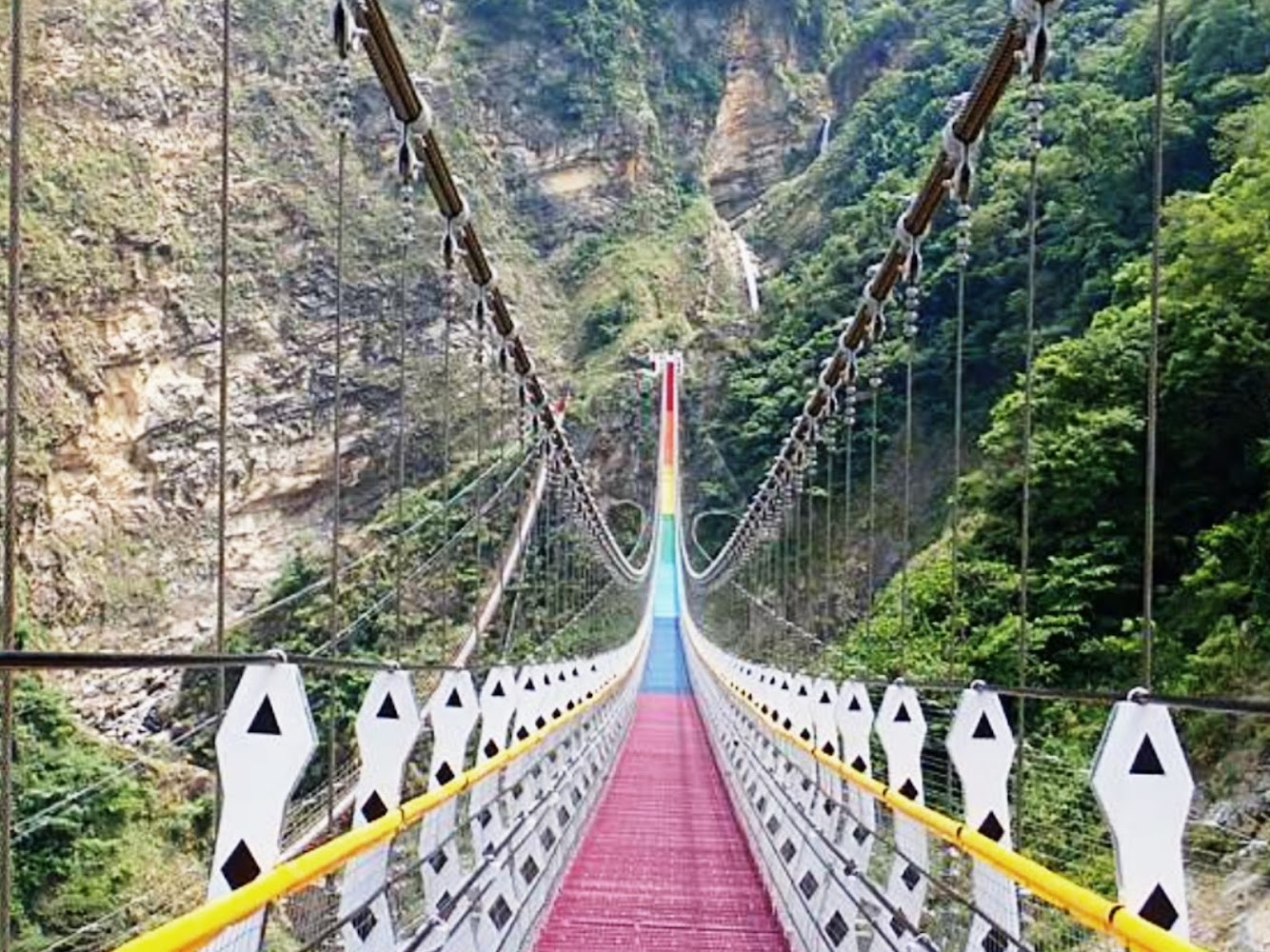 一座懸索吊橋,橋身色彩鮮豔,背景可見瀑布從山間流下。
