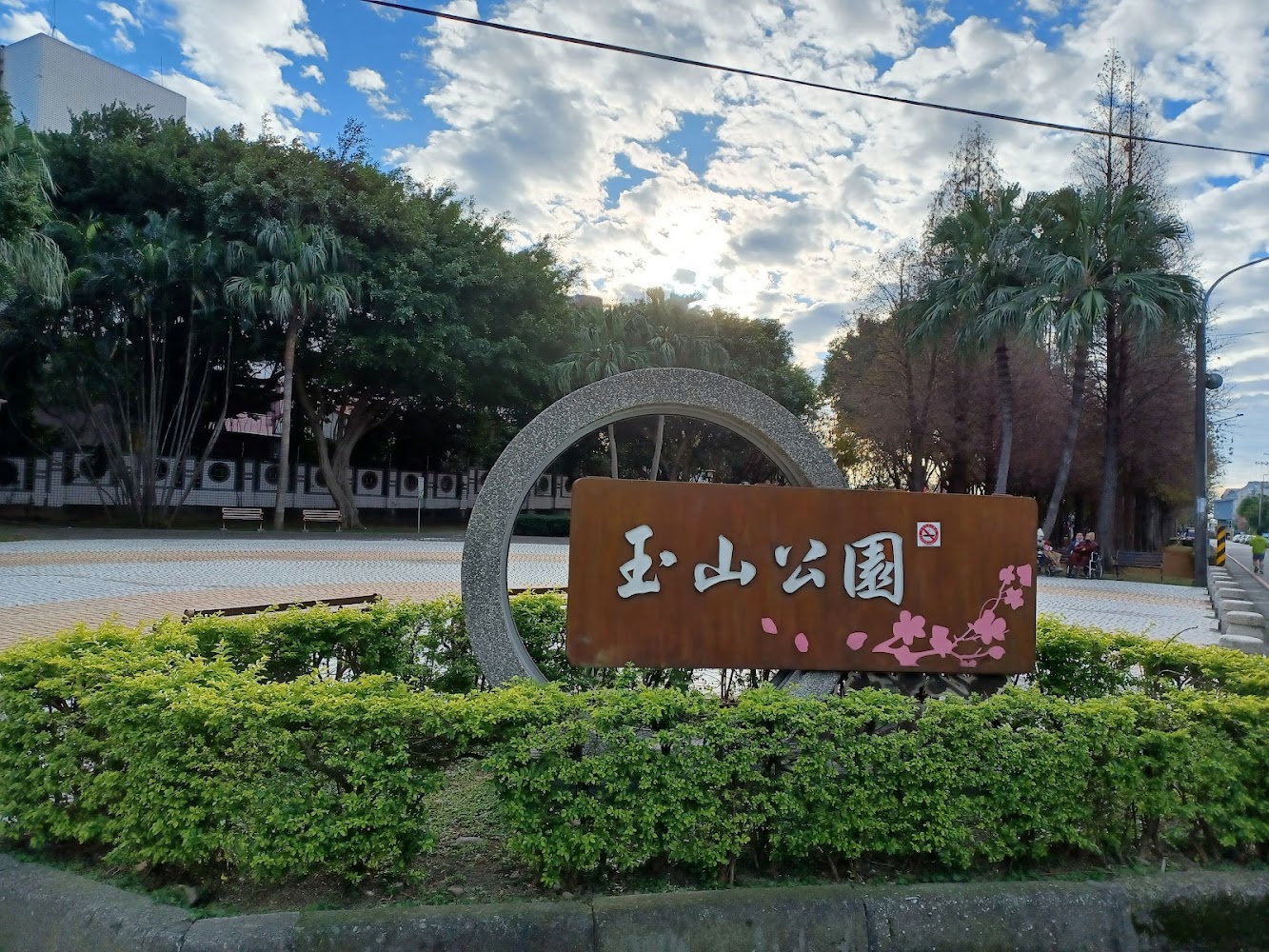 這張圖片主要展示了一個寫有『玉山公園』的大型公園指標牌，前方有綠色灌木，後方為茂密樹木和晴朗的天空。圖片無明顯浮水印。