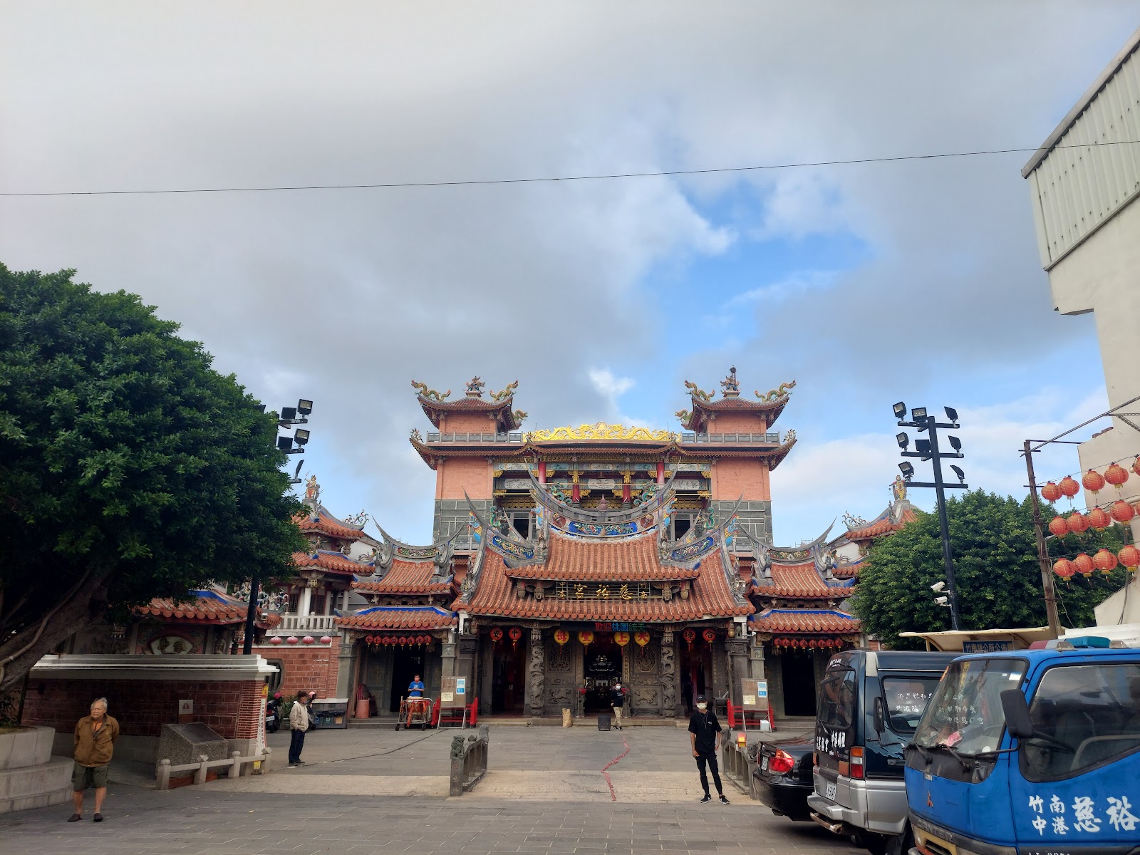 竹南中港慈裕宮正面外觀，廟宇屋頂飛簷精美，廣場有民眾走動，兩側有大樹
