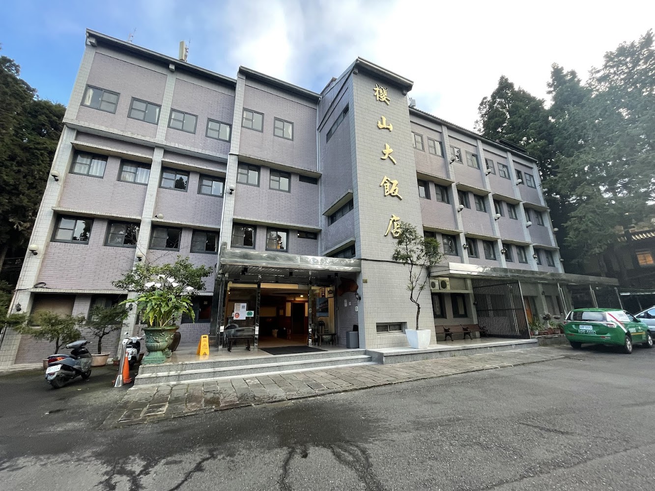 這張圖片主要是一棟飯店建築,外觀有明顯的中文招牌櫻山大飯店,四周有樹木、綠植、摩托車及計程車。
