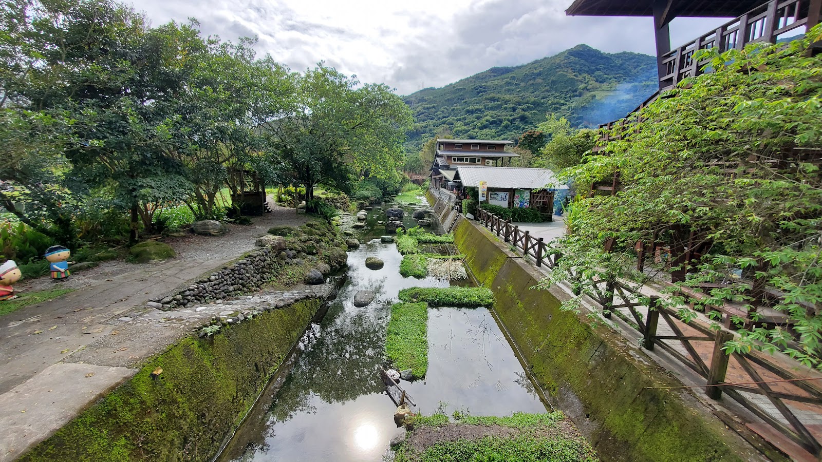 馬太鞍濕地溪流水道景觀，木欄杆棧道與青山背景，左側有原住民公仔裝飾