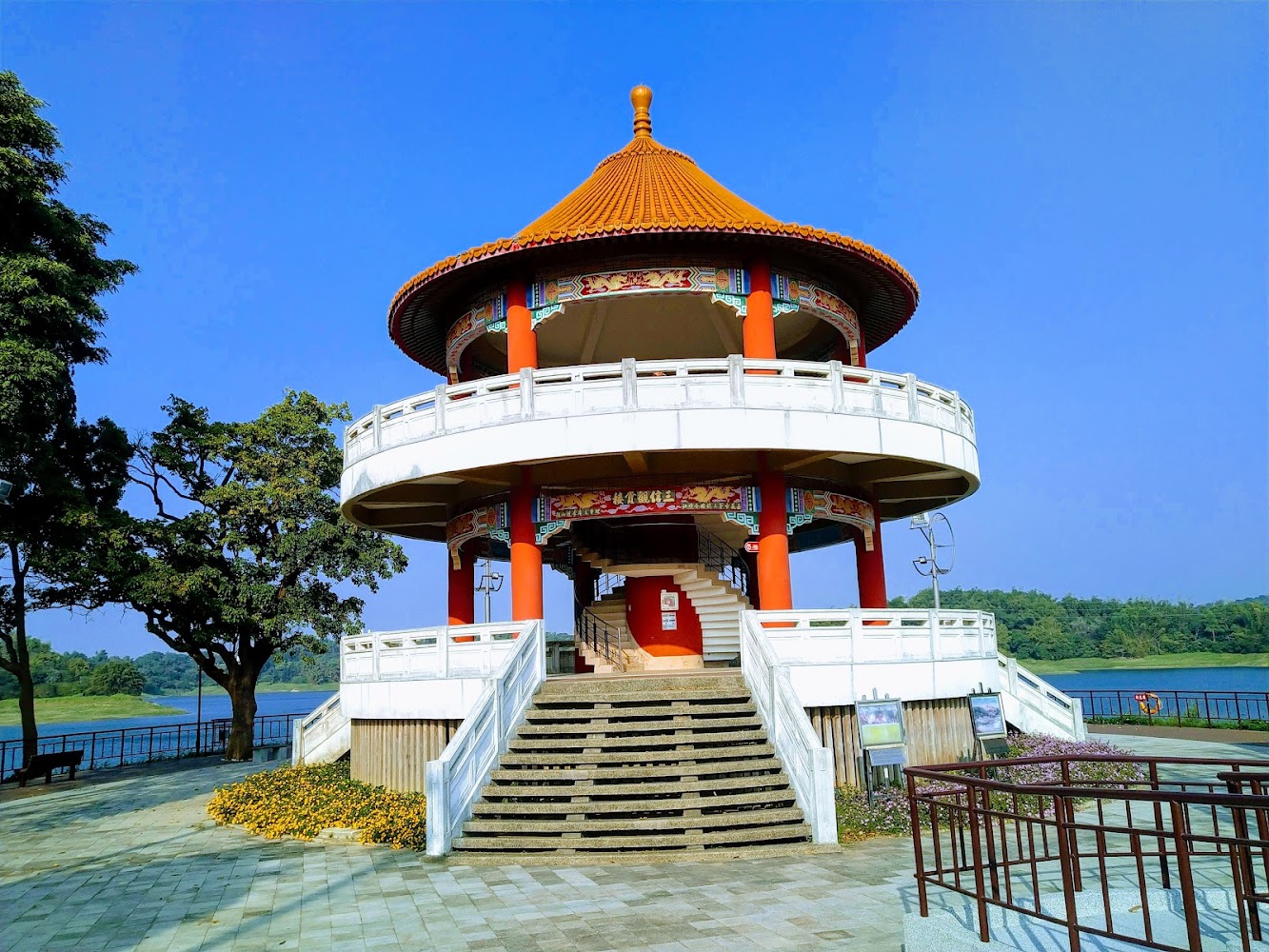 這張圖片主要是一座具有傳統中國風格的涼亭，圓形兩層建築位於湖邊，周圍有樹木和草地，背景是藍天白雲，整體環境清幽怡人。