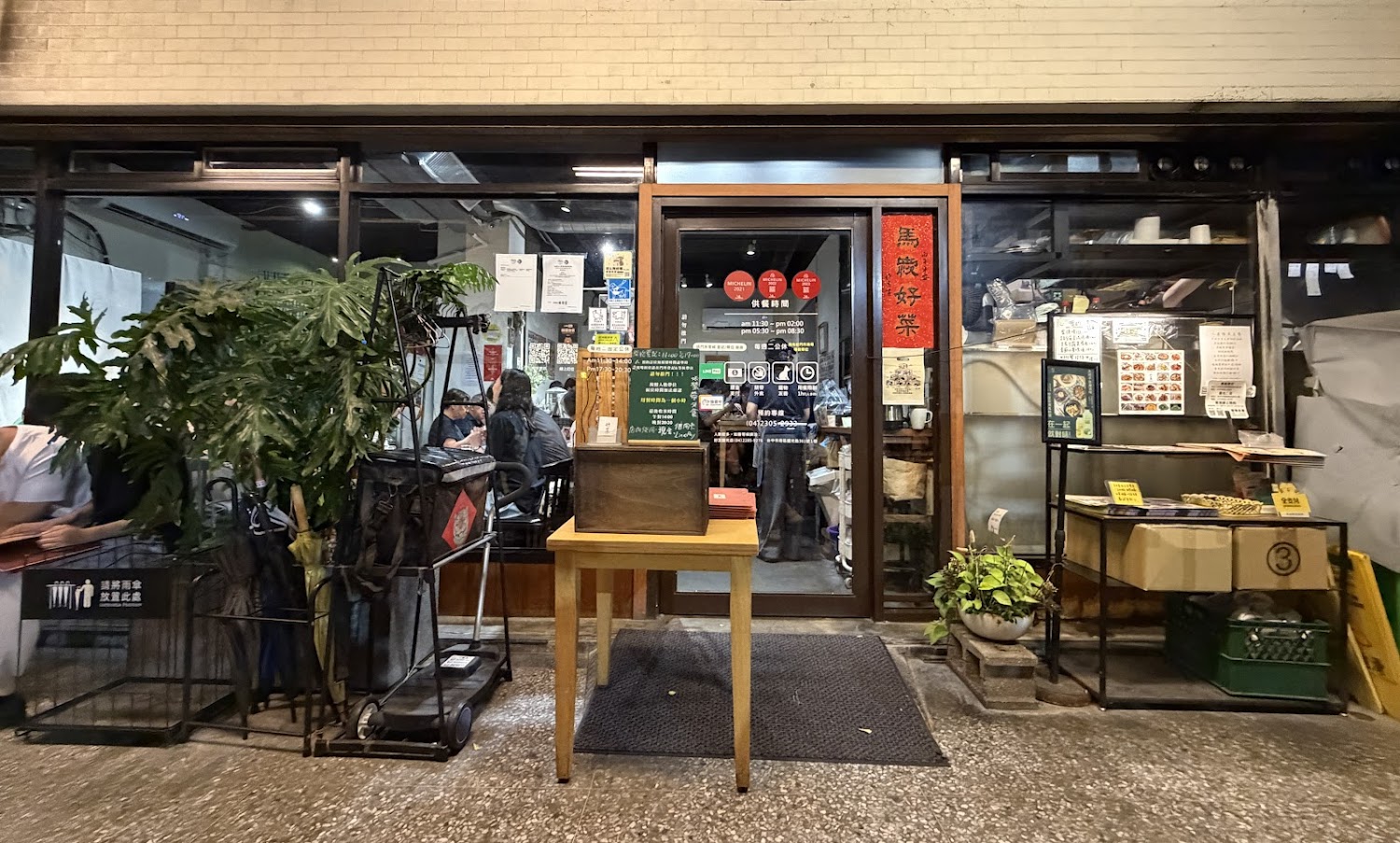這是一間餐廳門口，門上貼有營業時間及Michelin推薦標誌，旁邊有大葉植物和候位桌，店內可以看到用餐客人。圖片無明顯浮水印。