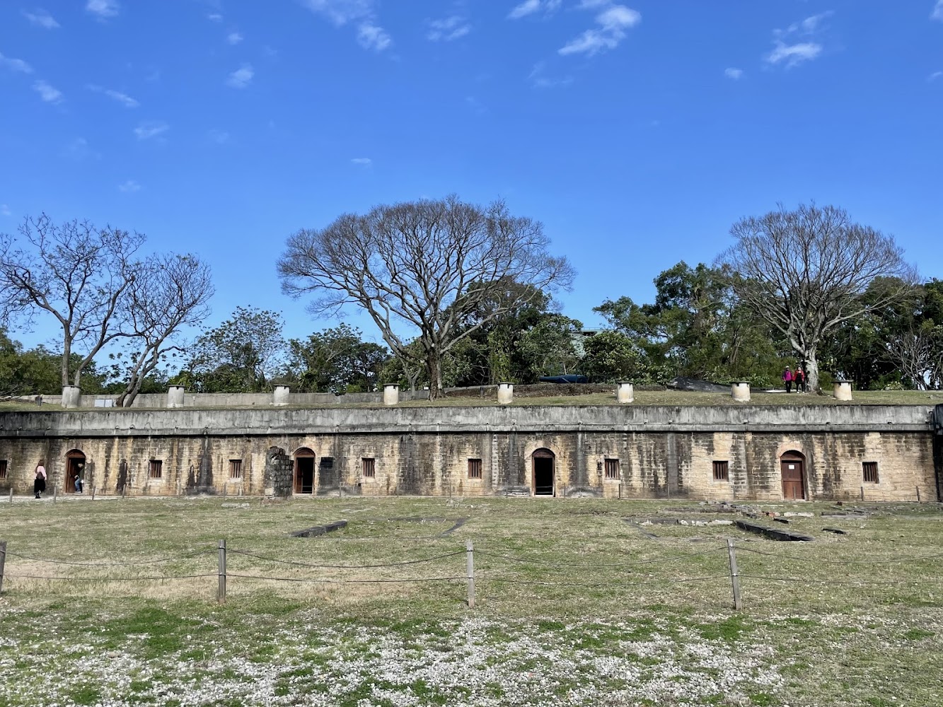 這張圖片展示一座歷史建築,外觀為石牆構造,門窗整齊排列,周圍有大片草地及幾棵樹。藍天與遊客點綴畫面,未見浮水印。