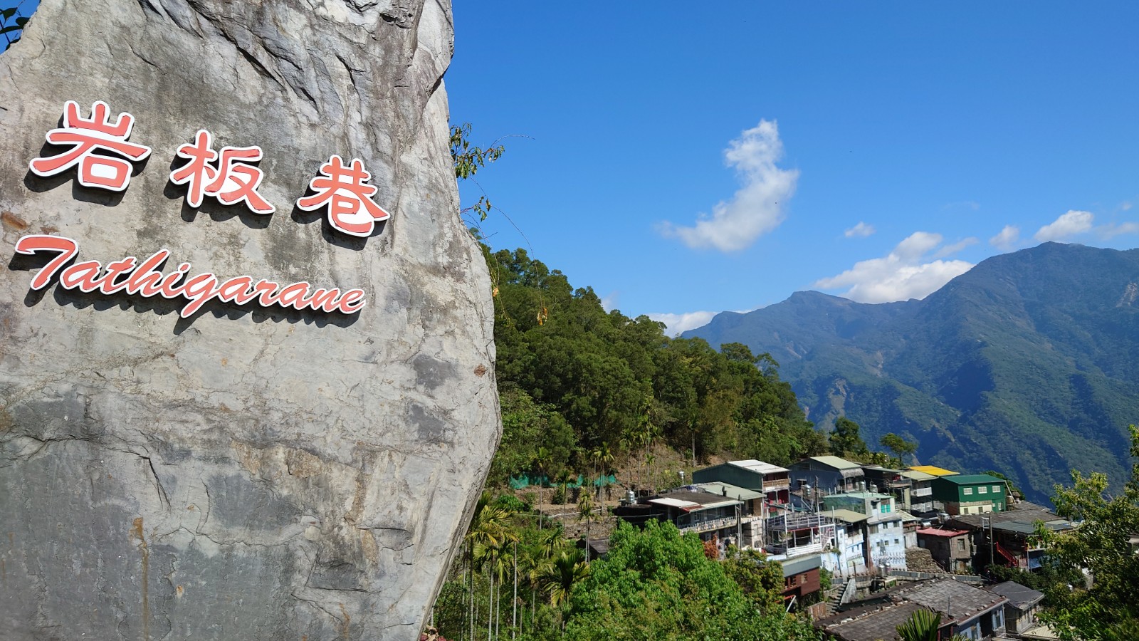 岩板巷石碑入口,刻有魯凱族語 Tathigarane,背景為山巒與聚落