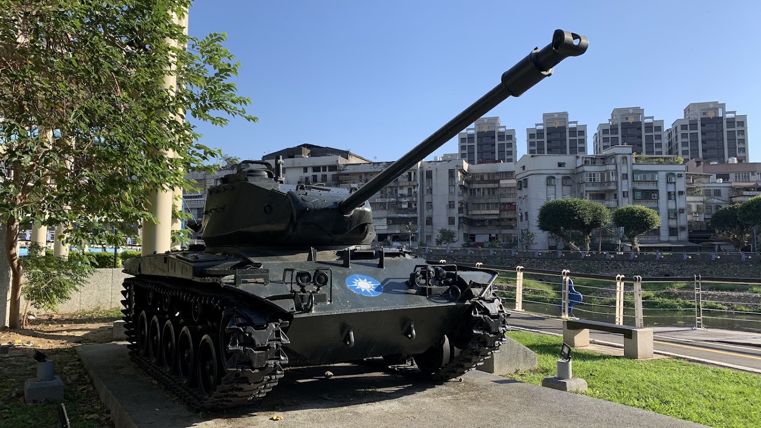 這張圖片中有一輛展示於戶外的戰車，戰車車身正面可見青天白日徽章，背景是城市建築和藍天，周圍有樹木和綠地。本圖片沒有明顯浮水印。