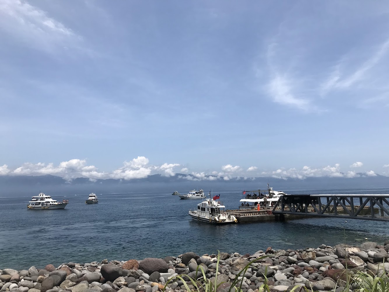 海岸邊的碼頭，有多艘船隻停泊或行駛於海面上，前景有圓石灘和草