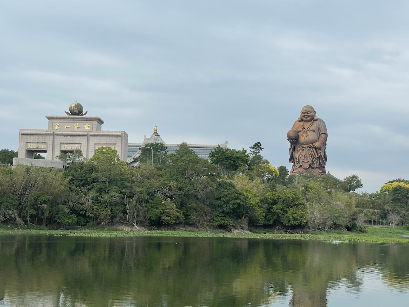 大型彌勒佛與寺廟建築前有樹林與水面