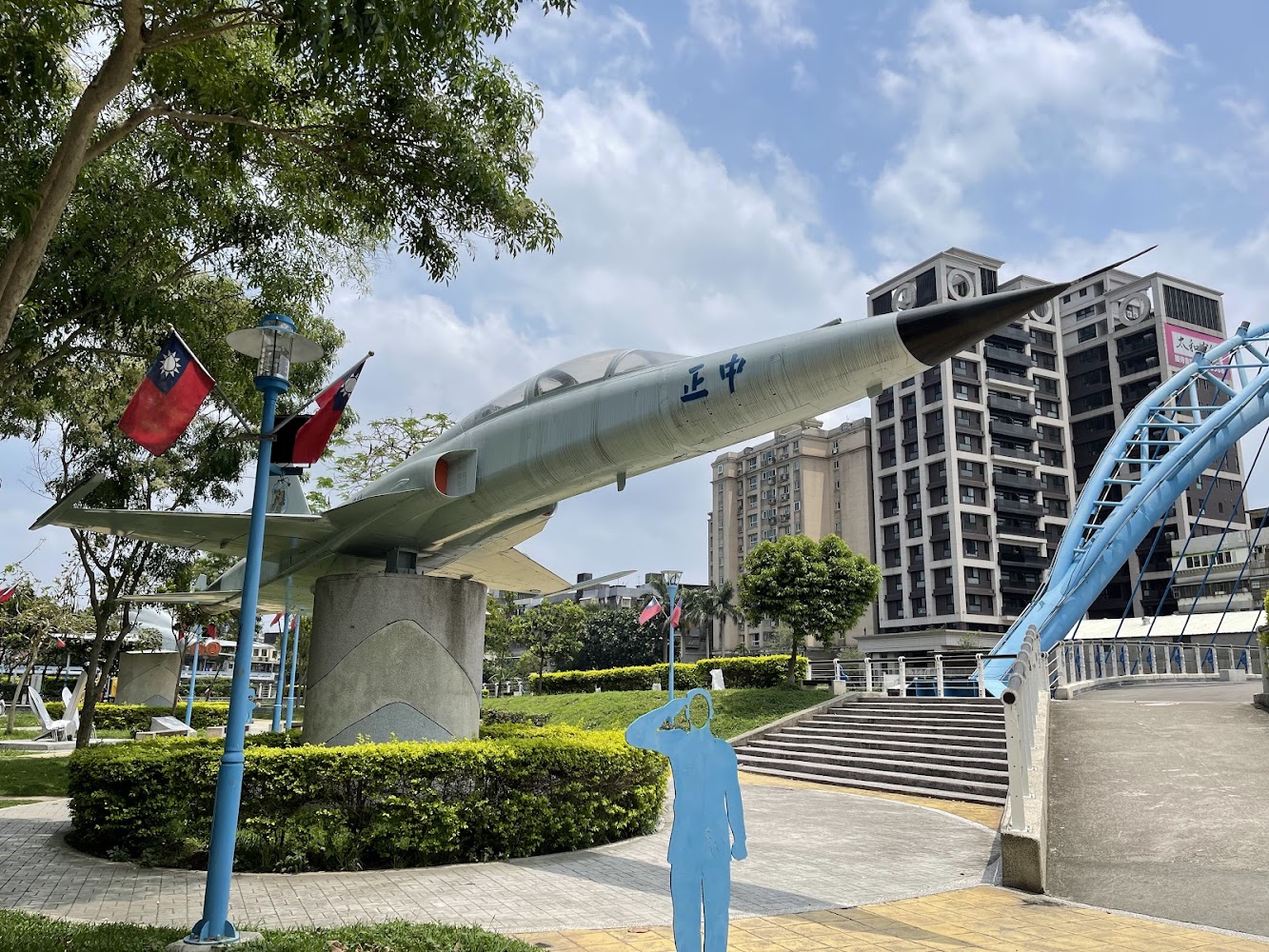 圖片中展示一架戰鬥機模型，機身上寫有“正中”二字。戰機架設於戶外公園，四周有台灣國旗、樹木、步道與高樓建築，還有一個藍色敬禮人形裝飾。圖片無明顯浮水印。