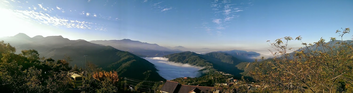 層層山巒以及山谷間雲海，前景中可見樹木房屋和黃色巴士