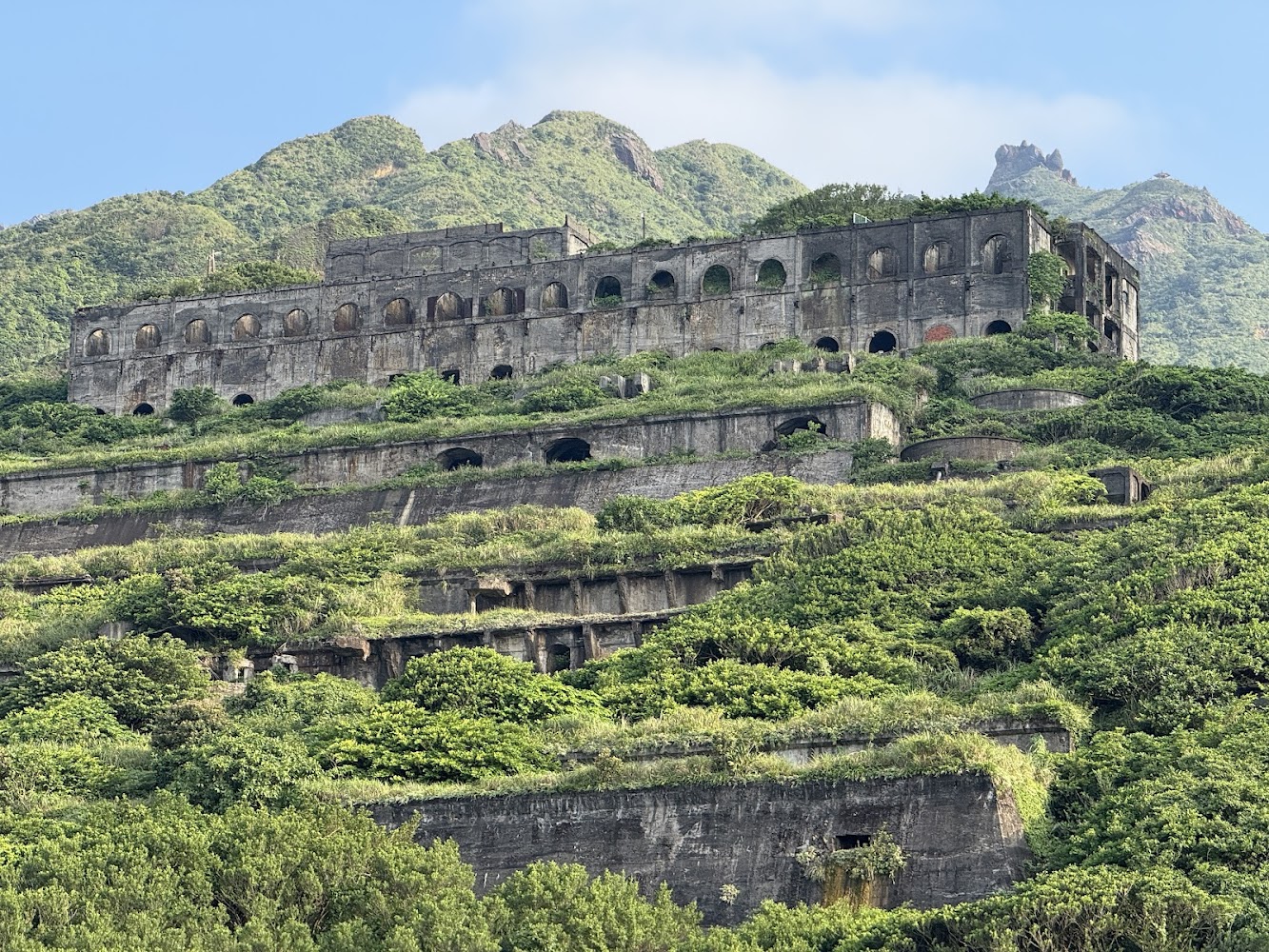 這張圖片主要是一座依山而建的廢棄建築遺跡,被濃密的綠植覆蓋,背景為青翠山巒。畫面未見浮水印。