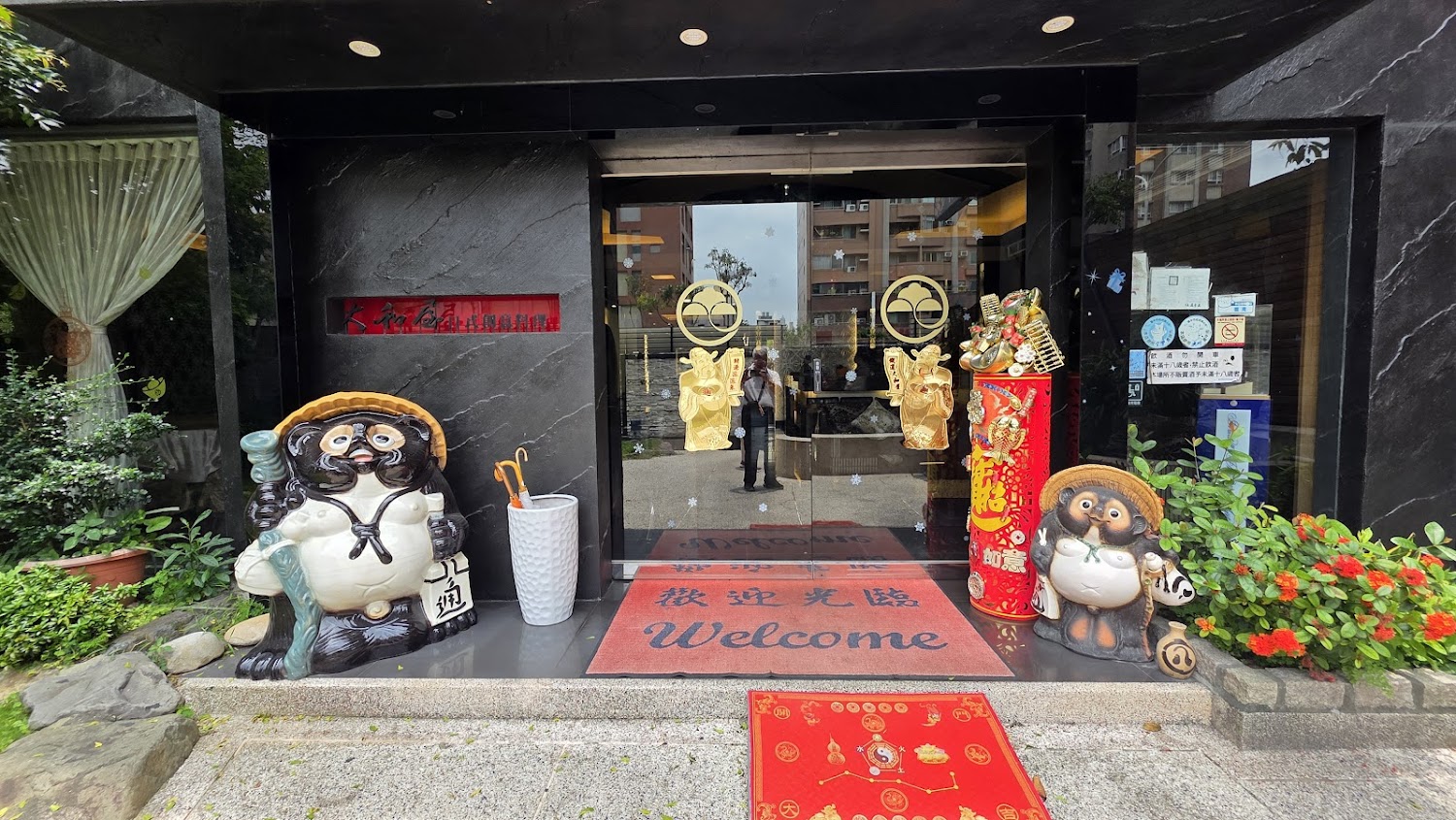這張圖片顯示一間餐廳入口，門口擺放兩尊狸貓擺飾，地上有紅色歡迎地墊與裝飾，兩側有植物，門上貼有金色裝飾與標誌，營造熱情且吉祥的氛圍。圖片右側貼有營業及防疫告示。圖片無明顯浮水印。
