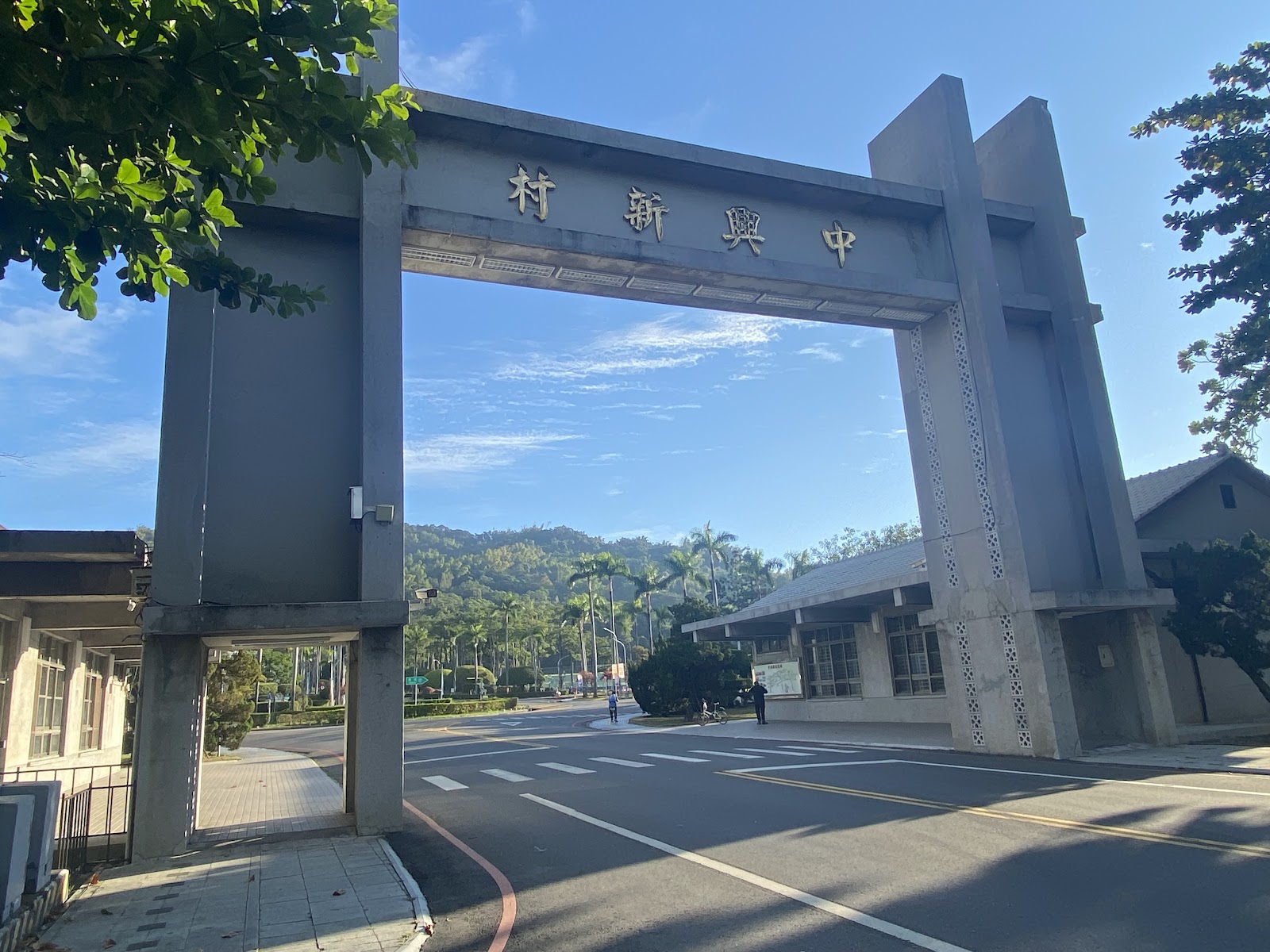 中興新村牌樓正面近景,灰色混凝土拱門刻有金字,兩側棕欄樹道