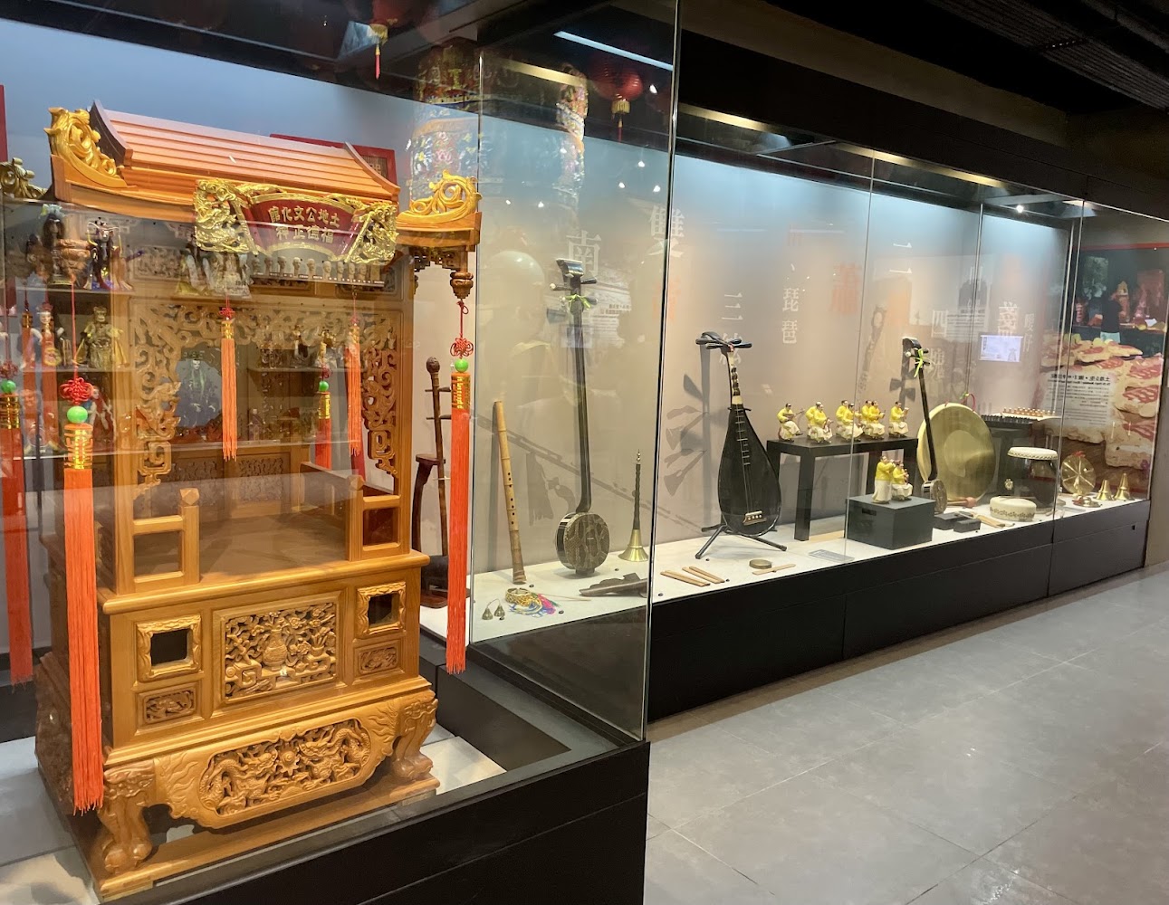 這張圖片展示了一個博物館展櫃，內有精緻的傳統木雕神轎以及各式民俗樂器，如琵琶、鑼、鼓等，反映出豐富的傳統文化特色。圖片無明顯浮水印。