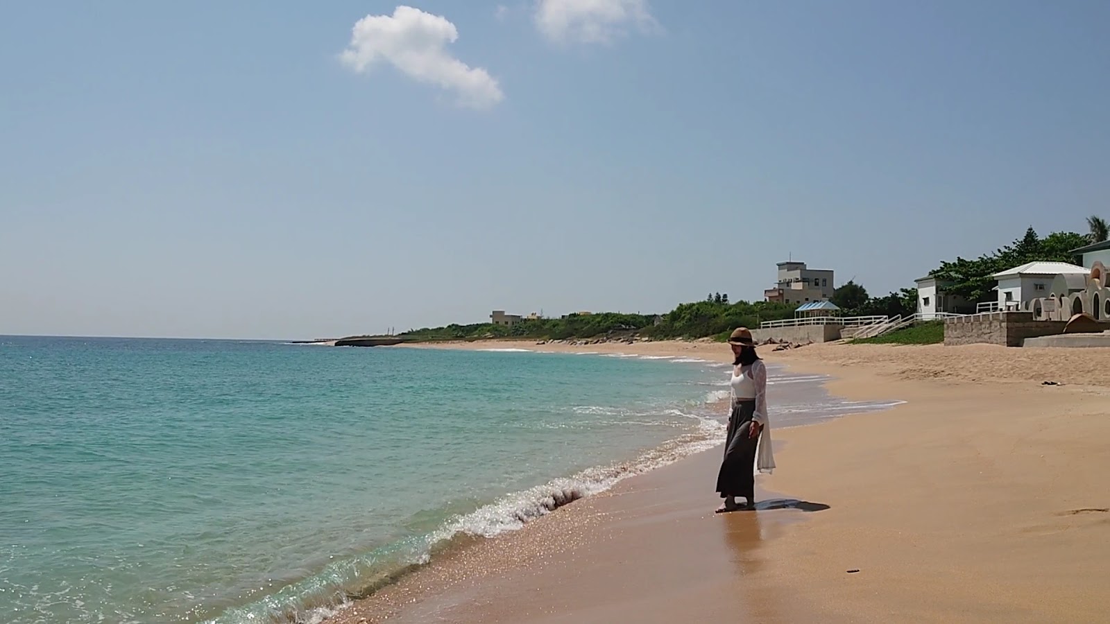 網垵口沙灘，戴帽女士佇立碧藍海水旁，遠景有度假建築