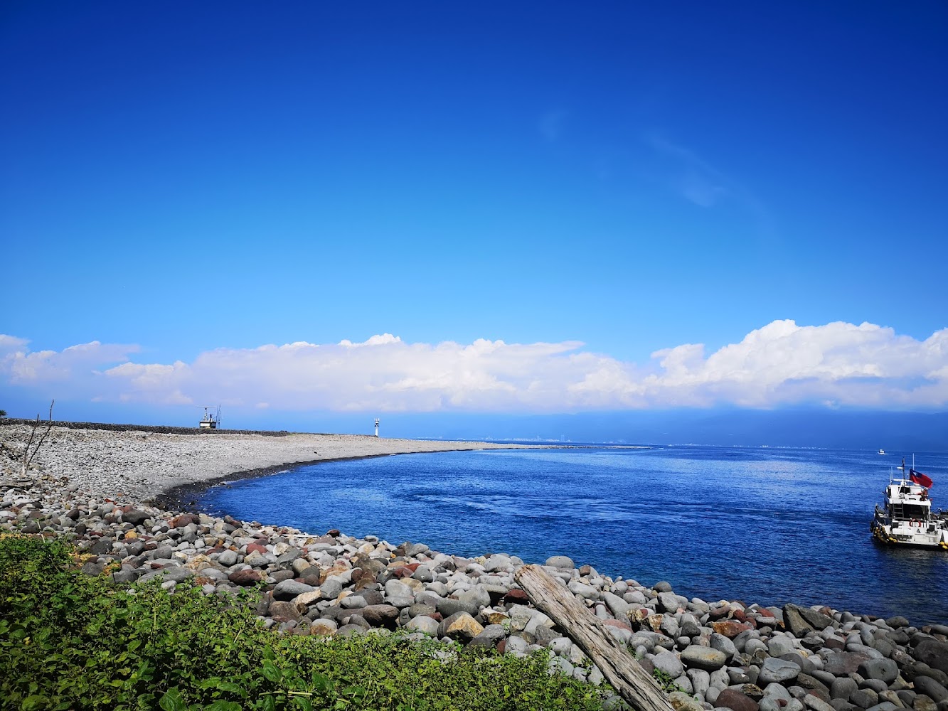 礫石海灘與蔚藍海水，遠處可見燈塔和小船，天空晴朗