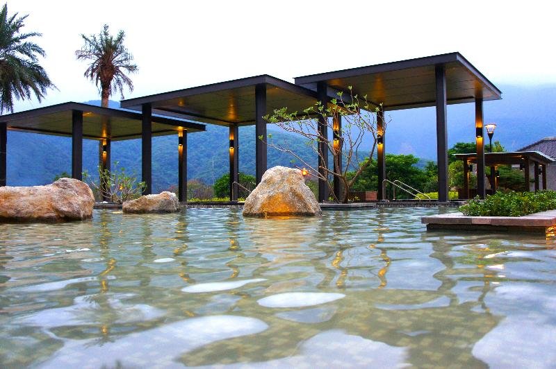 戶外溫泉池,池中有大石與水面波紋,背景為現代涼亭建築和山景