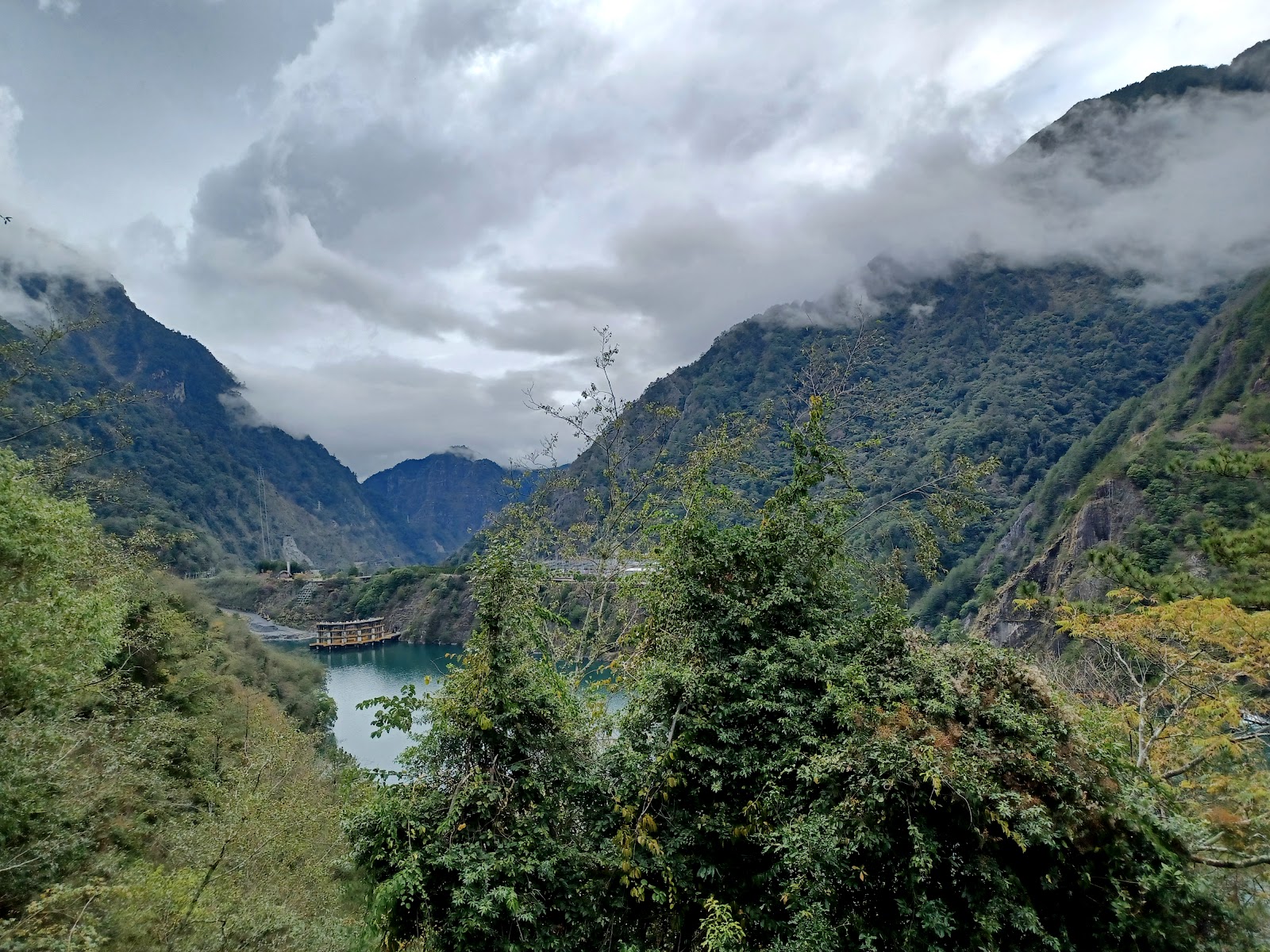 穿越山坡樹叢俯瞰德基水庫全景，可見水壩建築與壯闊雲層