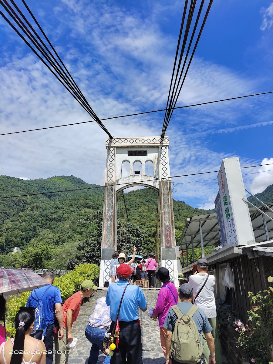 武界吊橋橋頭白色拱門正面,遊客排隊準備走上吊橋,背後是藍天與青山