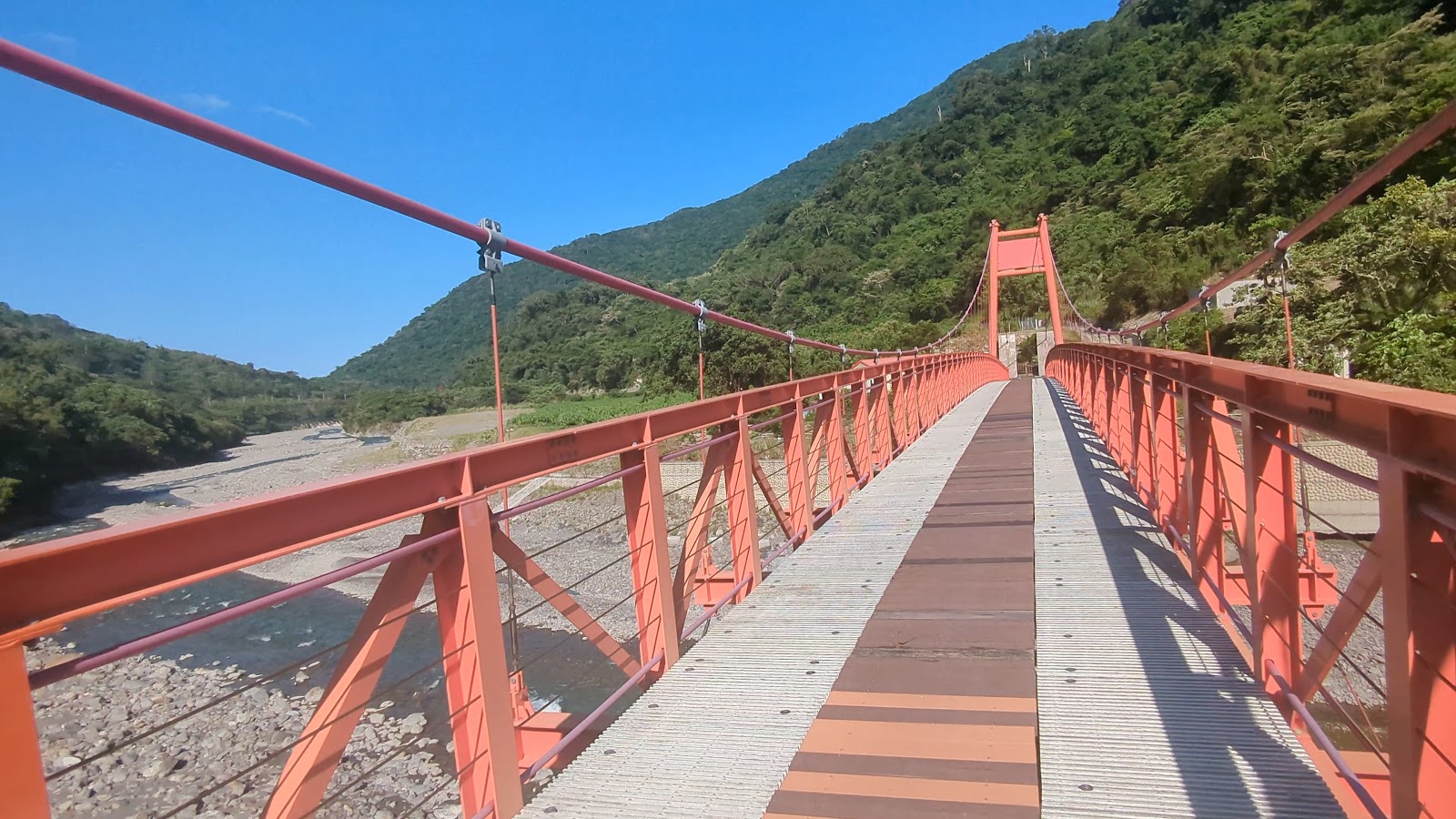 站在魯拉克斯吊橋橋面往前透視,橙紅橋欄延伸,橋下礫石河床,藍天晴朗