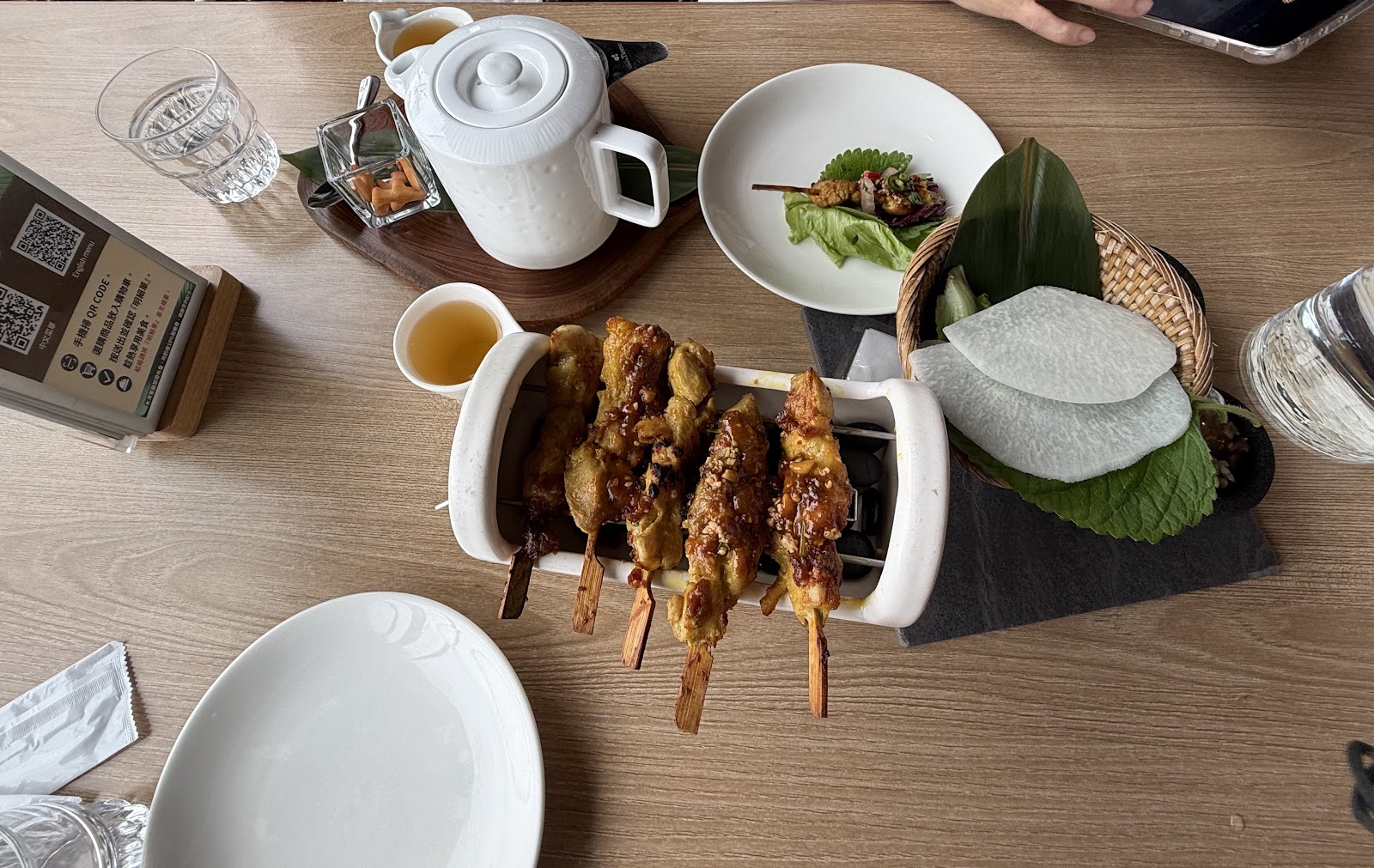 水灣餐廳東南亞風味料理俯拍,沙嗲串與前菜擺放在木桌上,旁邊搭配白色茶壺