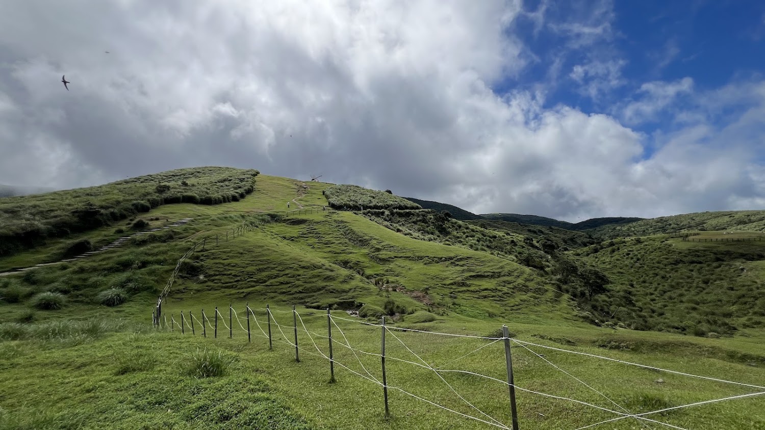 青翠山丘草原與層層雲朵,前景有欄杆