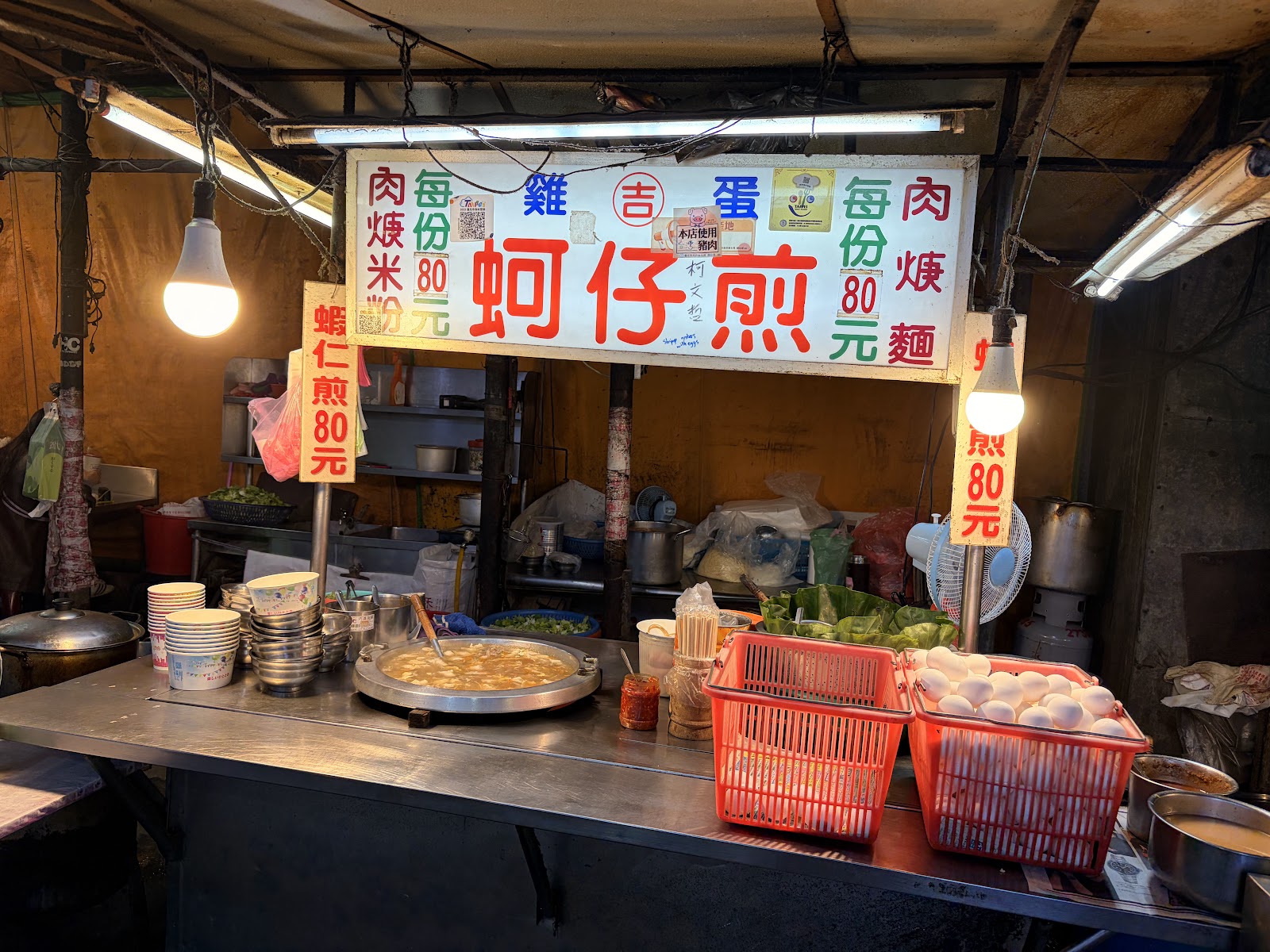 吉雞蛋蚵仔煎攤位招牌，黃底寫著蚵仔煎每份80元，鐵板與雞蛋籃陳列在攤車上
