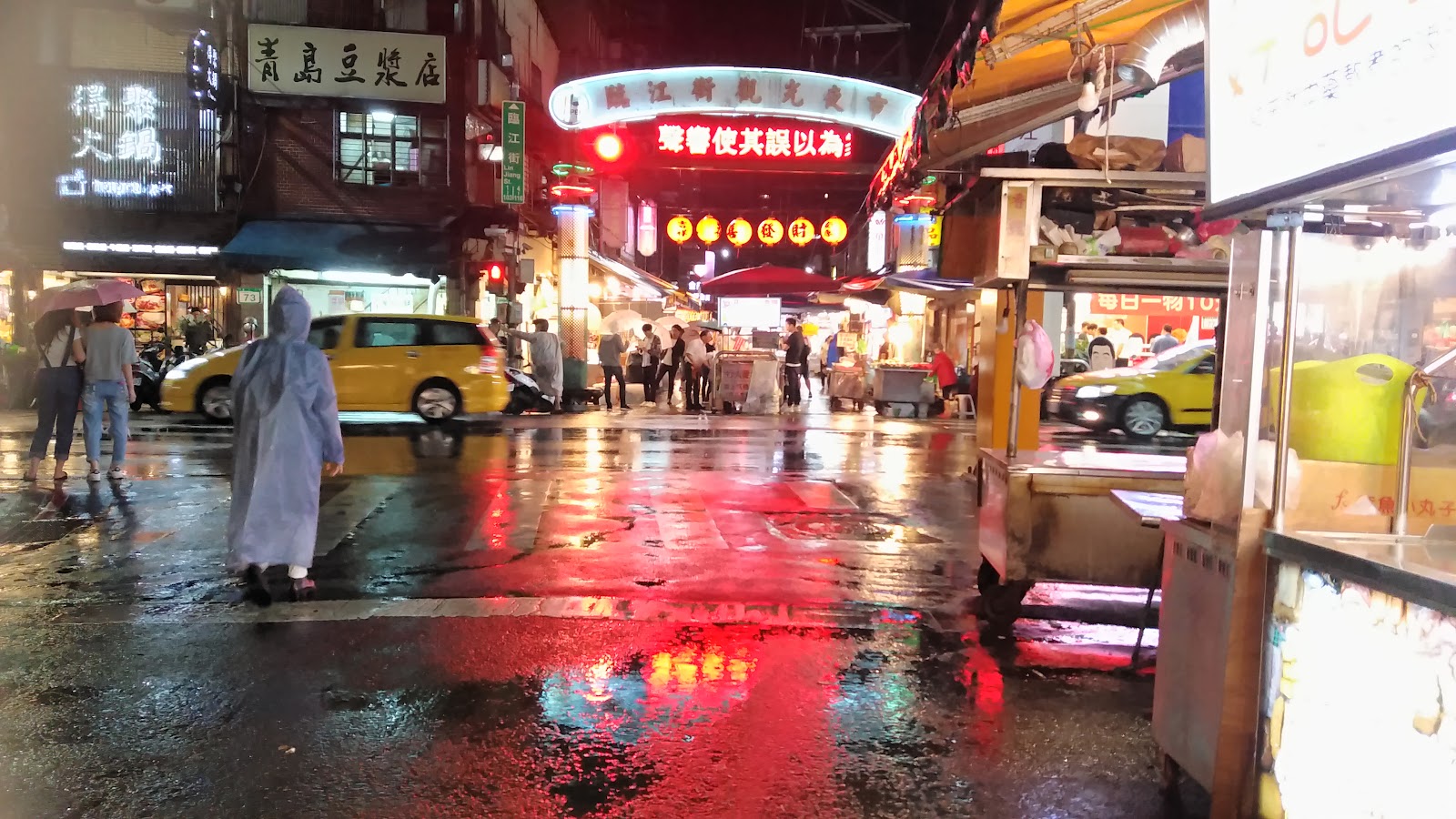 臨江街觀光夜市雨夜街景，攤車與招牌霓虹燈倒映濕滑地面，通化夜市夜間氛圍