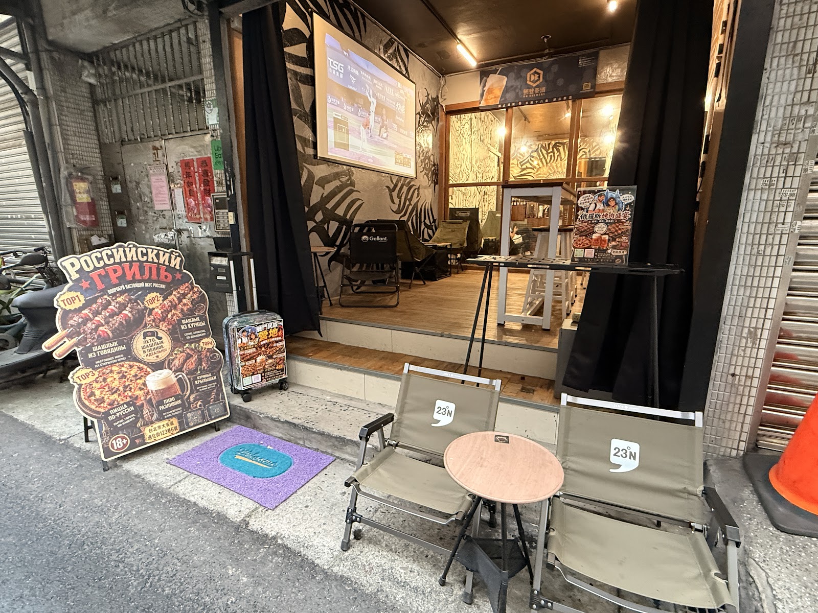 俄羅斯烤肉露營咖啡廳店門口，擺放露營折疊椅與小圓桌，俄文招牌與烤肉海報