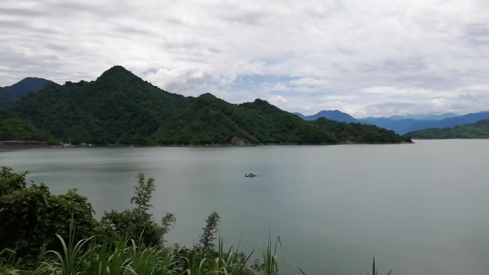 曾文水庫全景遠景，湖面灰綠色，前景青翠灌木，遠處一艘小船，背景群山連綿