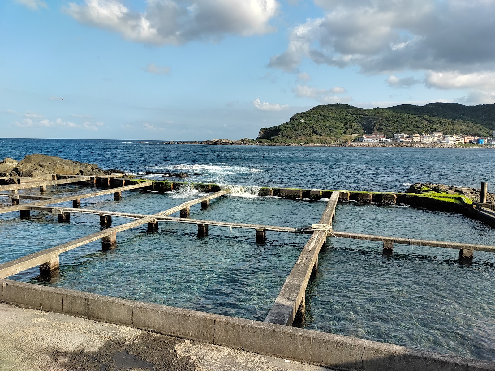 貢寮海岸石造格狀魚塭，海浪湧入方格石池，背景貢寮灣翠綠山丘與小鎮