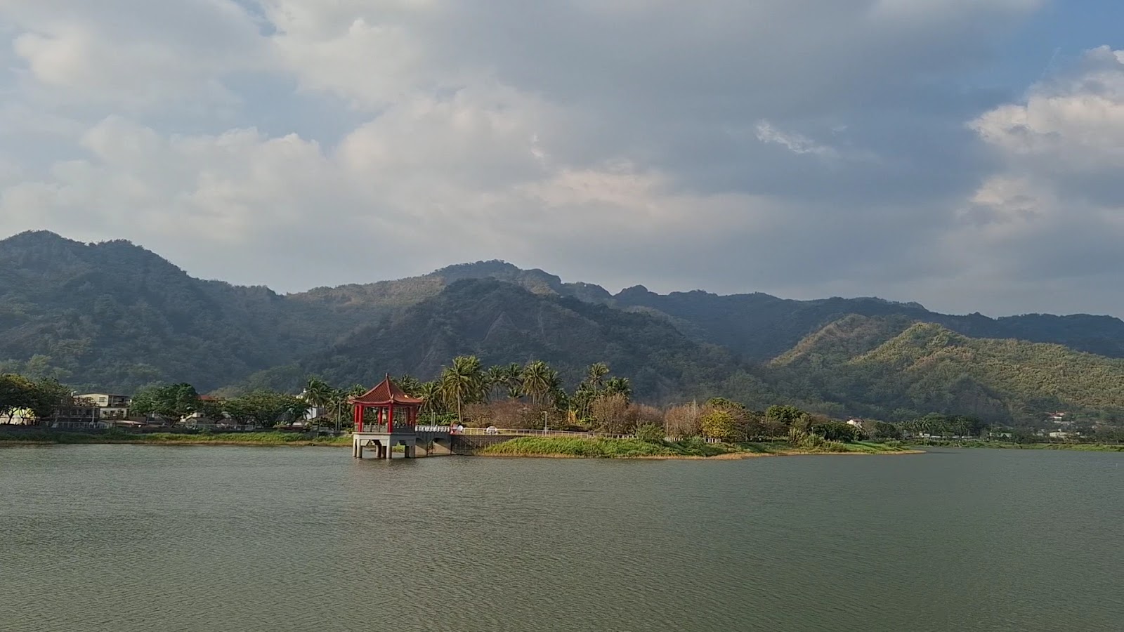 藍天白雲下的中圳埤全景,白色拱橋延伸至湖心紅亭,遠山環繞