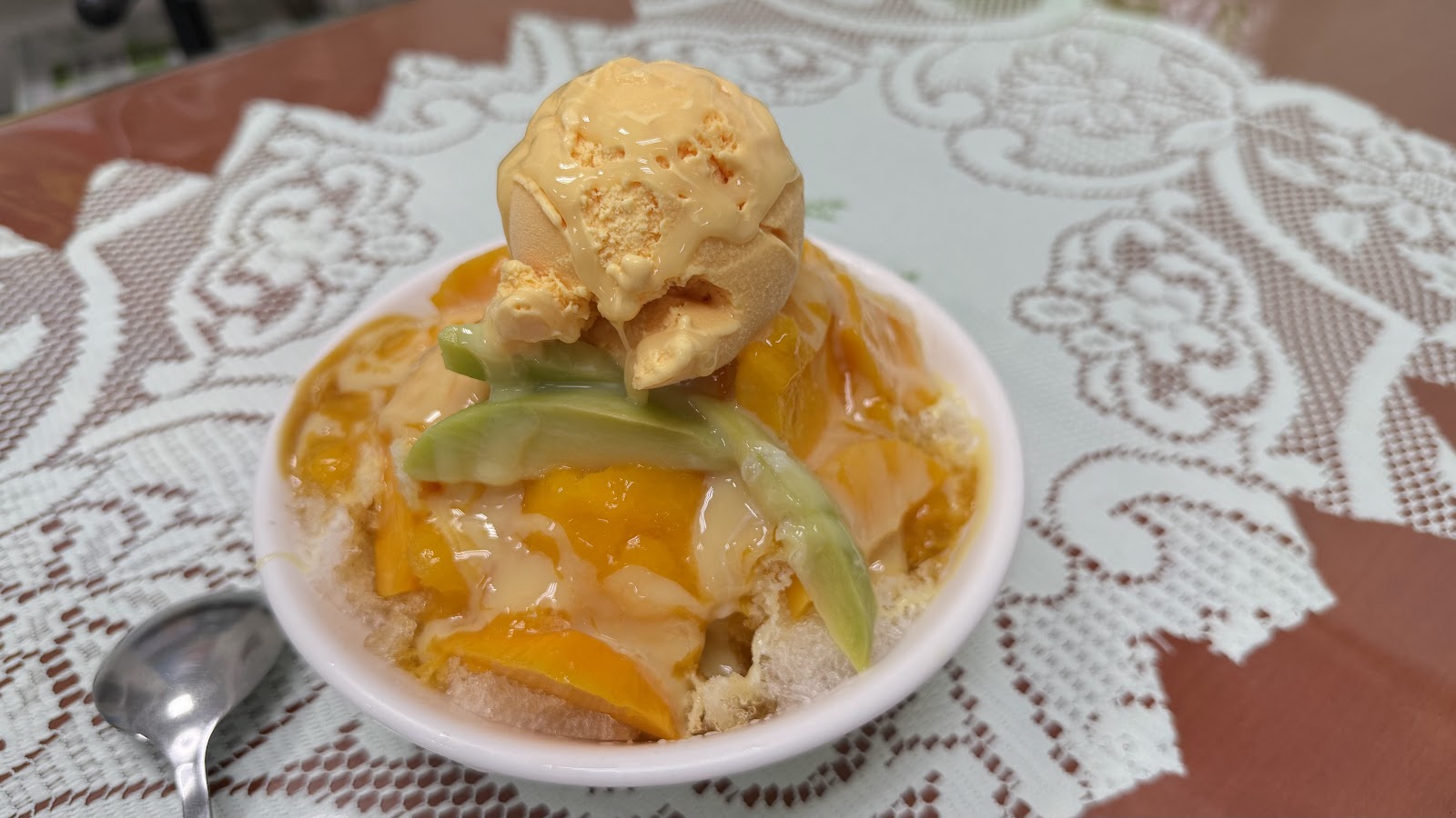 玉梅芒果冰的芒果冰淇淋球特寫，橙黃色冰淇淋放在碗中，花卉圖案桌巾背景