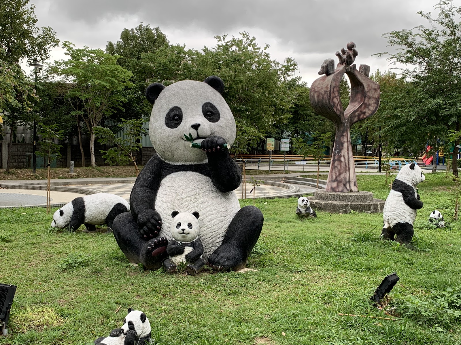楓樹河濱公園大型熊貓裝置藝術,草坪上一大一小熊貓雕像,旁邊有多隻小熊貓散落,背景有兒童遊樂設施